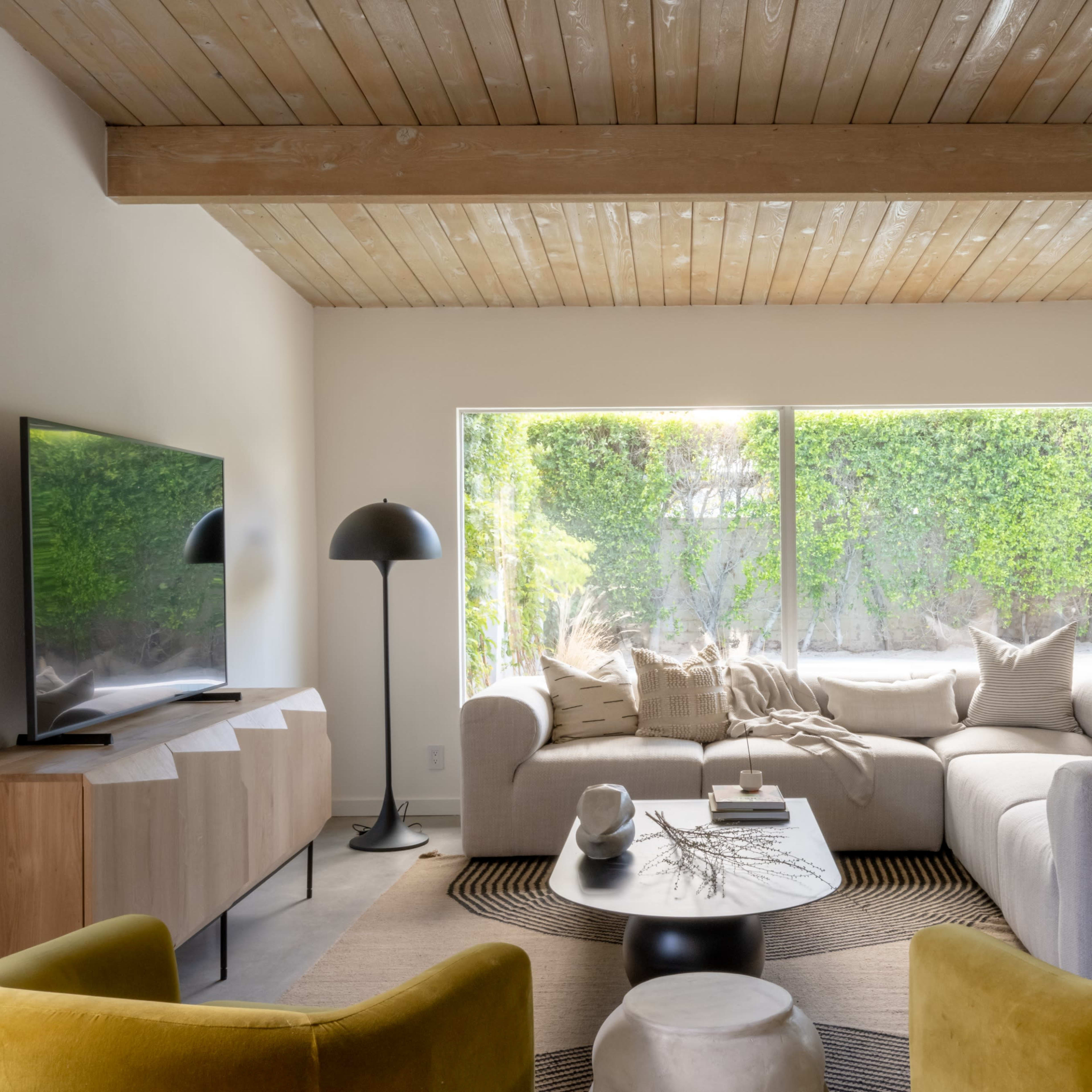 A modern living room features a light-colored sectional sofa, a round coffee table, and a large window overlooking a green outdoor space.