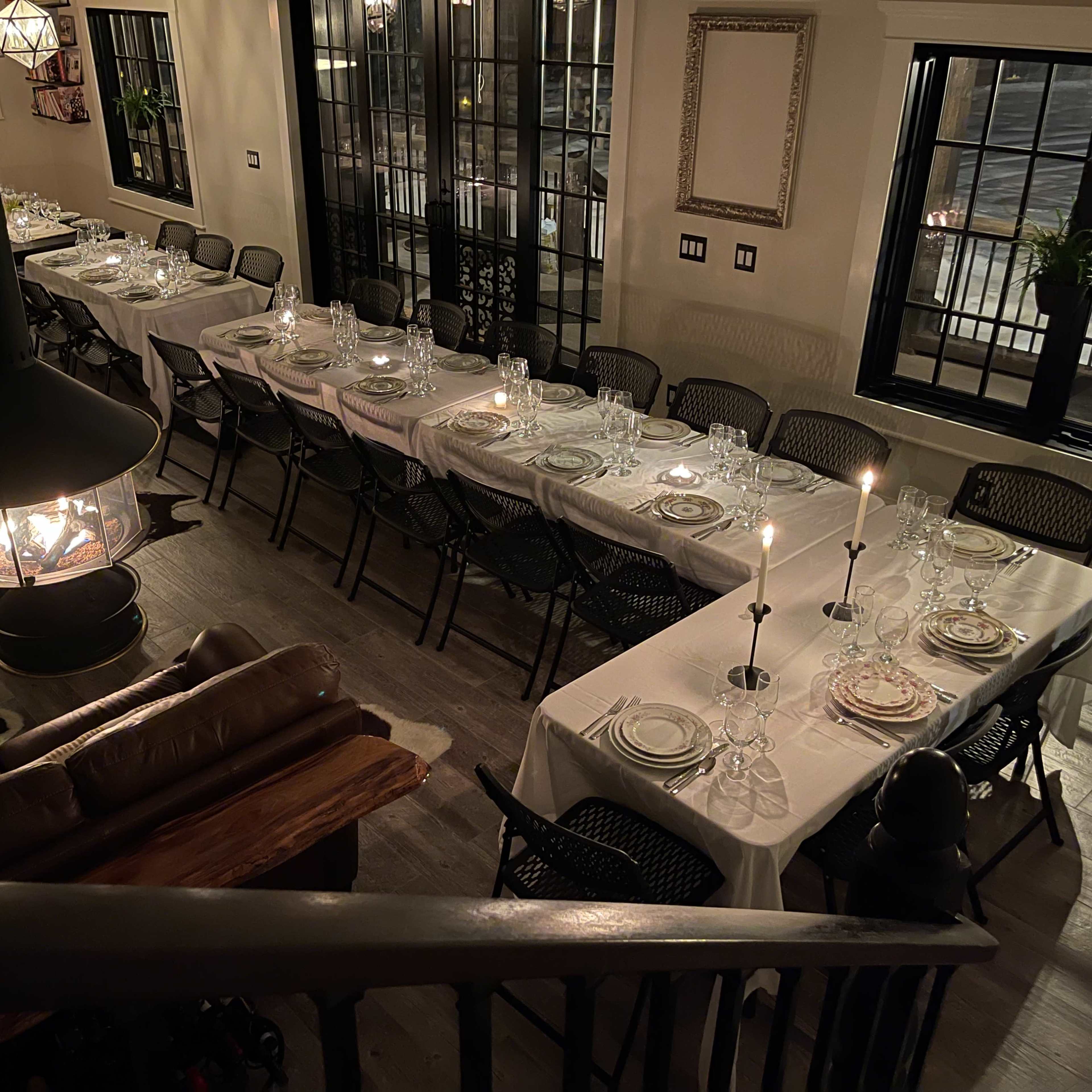 A spacious dining area is set with multiple tables, each covered with white tablecloths and arranged with plates and candles, illuminated by soft lighting.