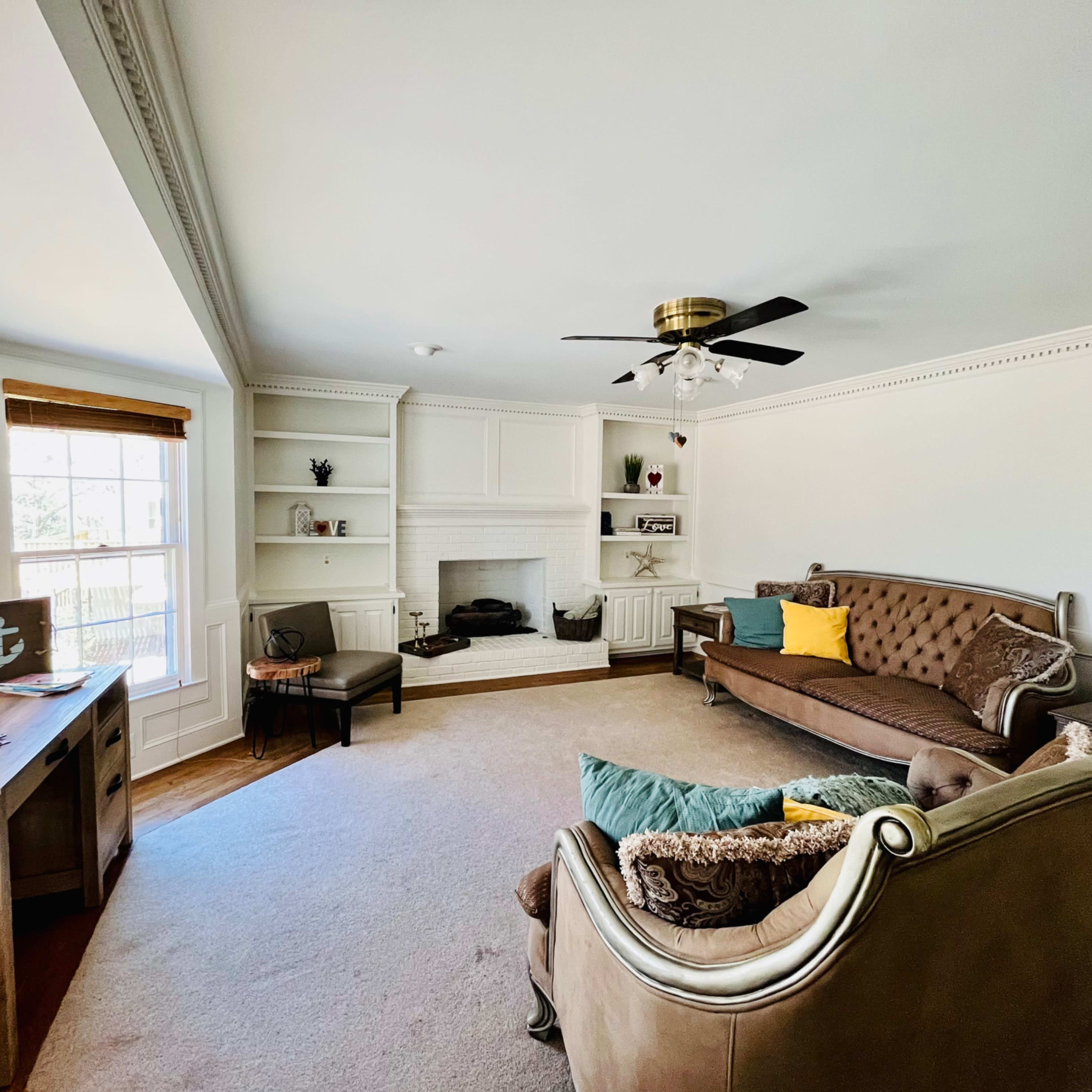 A living room featuring a brown sofa, two armchairs, a fireplace, built-in shelves, and large windows with wooden blinds.