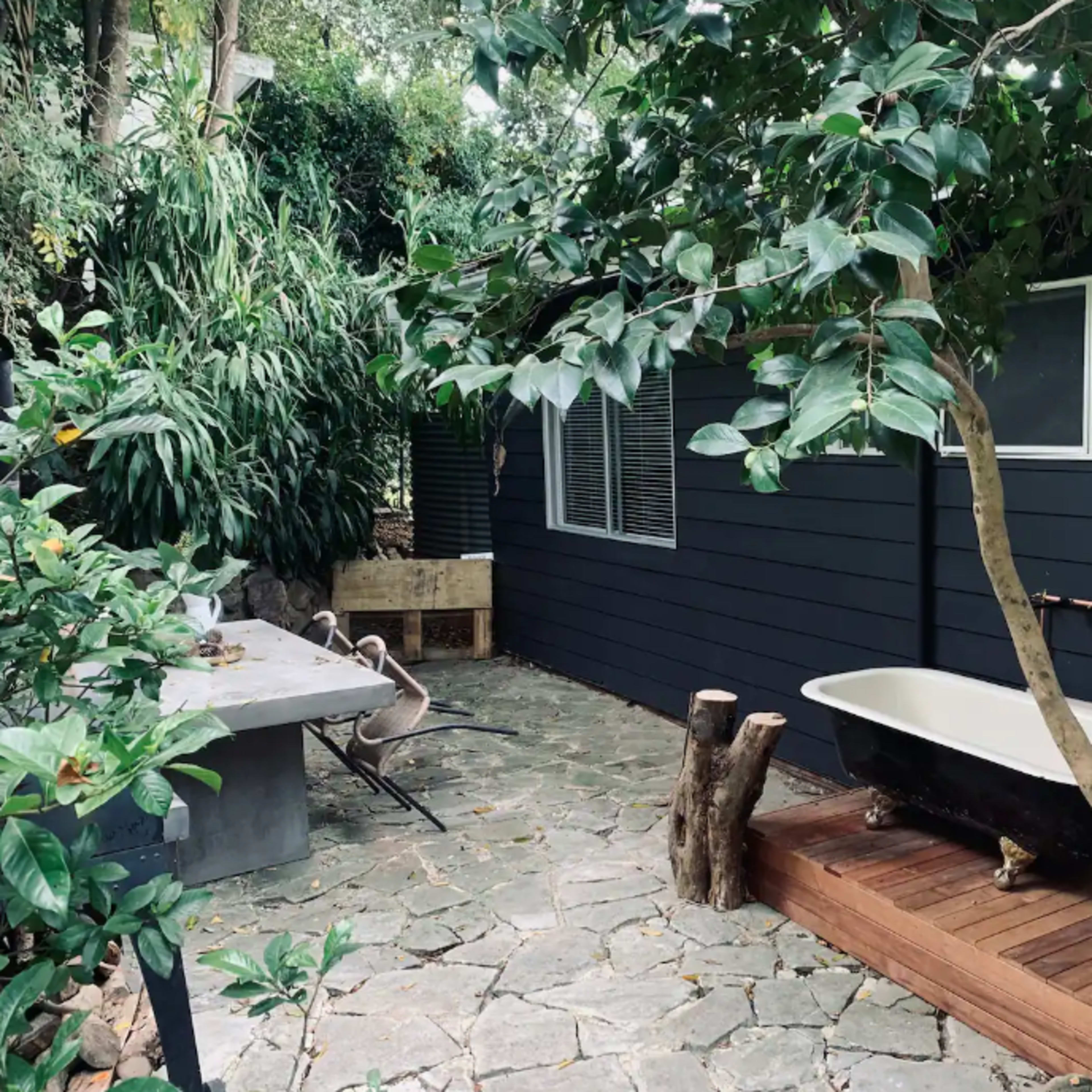 A stone patio features a freestanding bathtub surrounded by trees and plants, next to a wooden deck and a dining area.