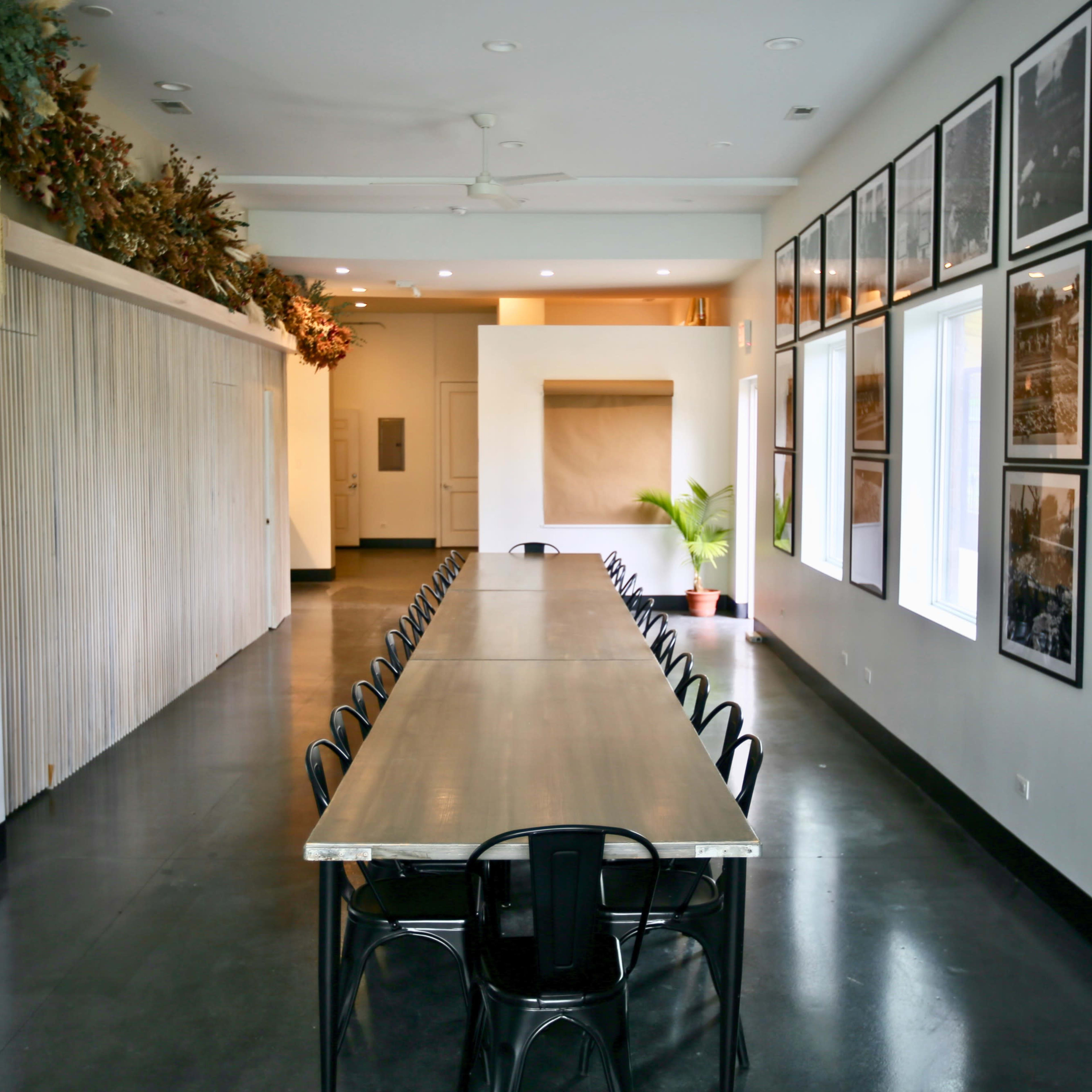 A long, rectangular dining table is placed in a spacious room with large windows and black flooring, surrounded by framed photographs on the walls.