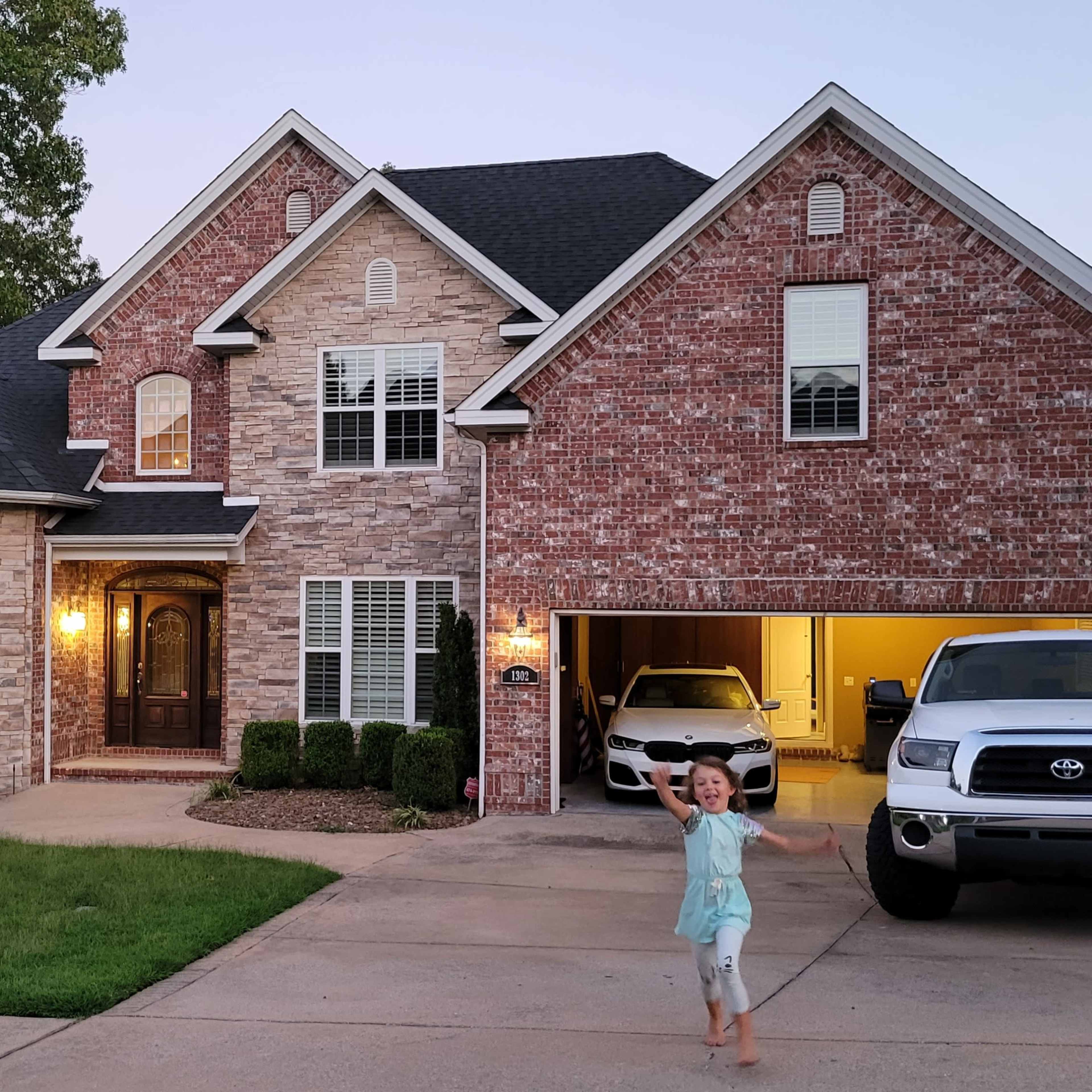 A child runs toward the camera in front of a large brick house with a neatly landscaped yard and a parked truck in the driveway.
