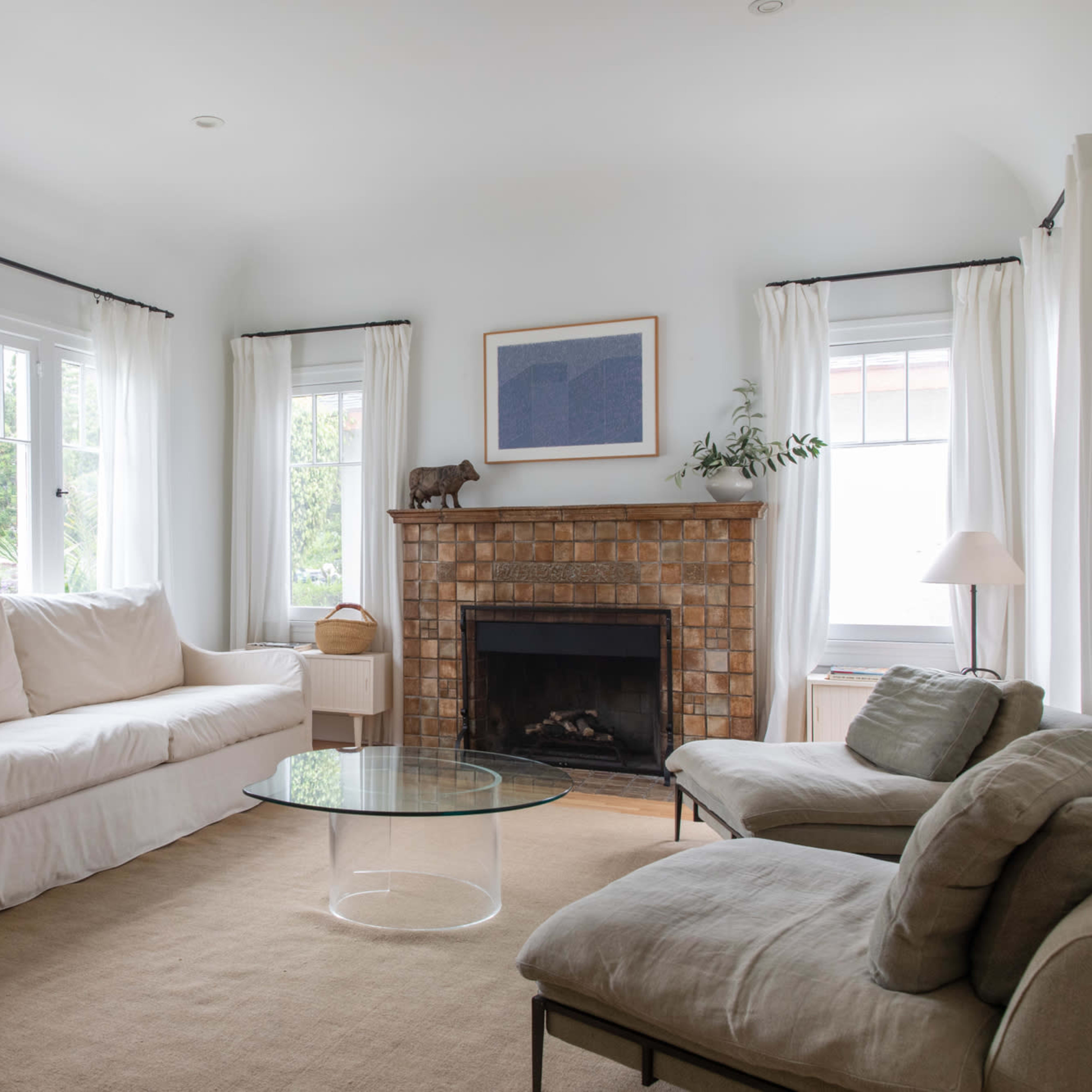 The living room features a white sofa, two light-colored armchairs, a glass coffee table, and a brick fireplace, all illuminated by natural light through large windows.