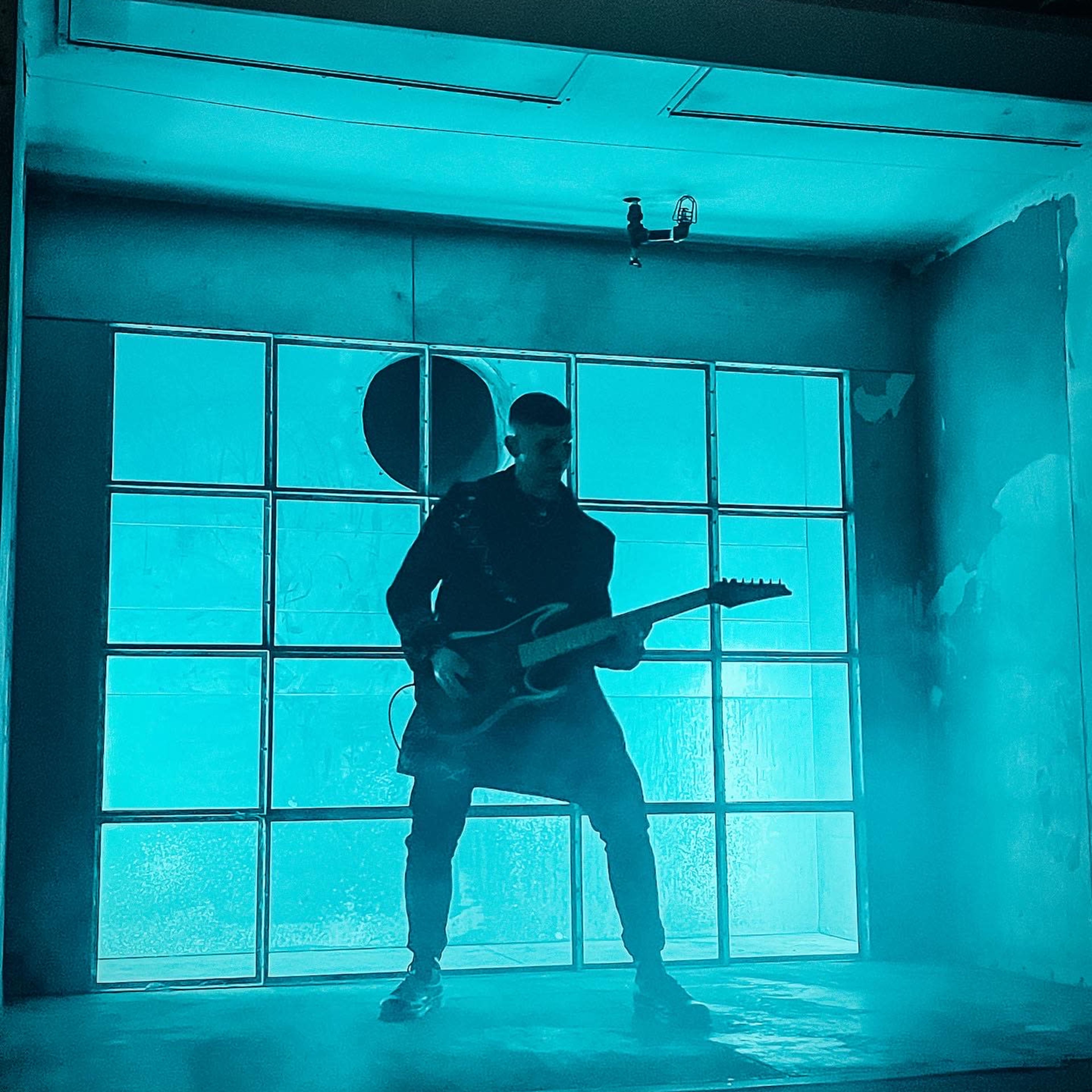 A musician plays an electric guitar in front of a large, illuminated window with a smoky backdrop.