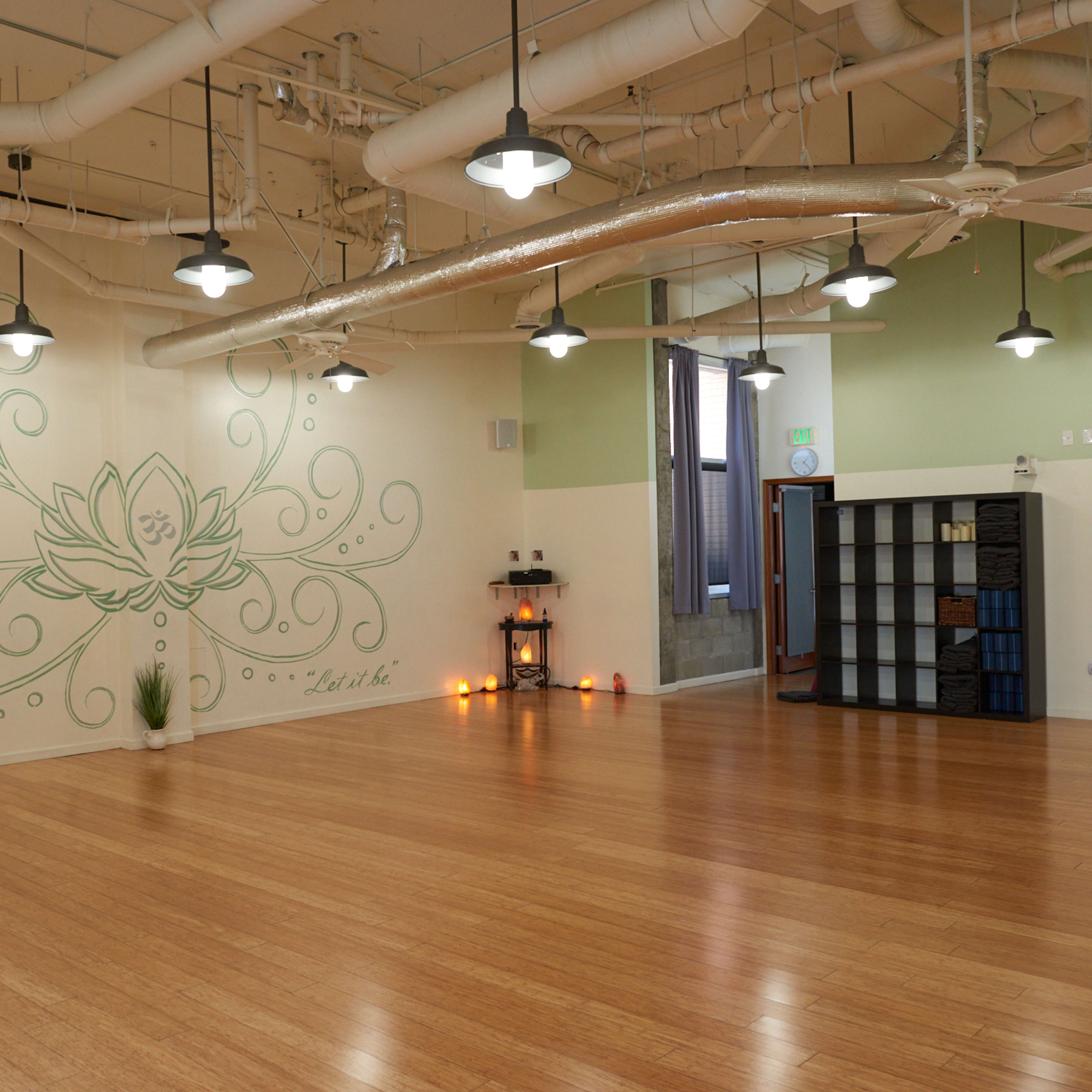 A spacious yoga studio with bamboo flooring, a green wall mural of a lotus flower, and various decorative elements.