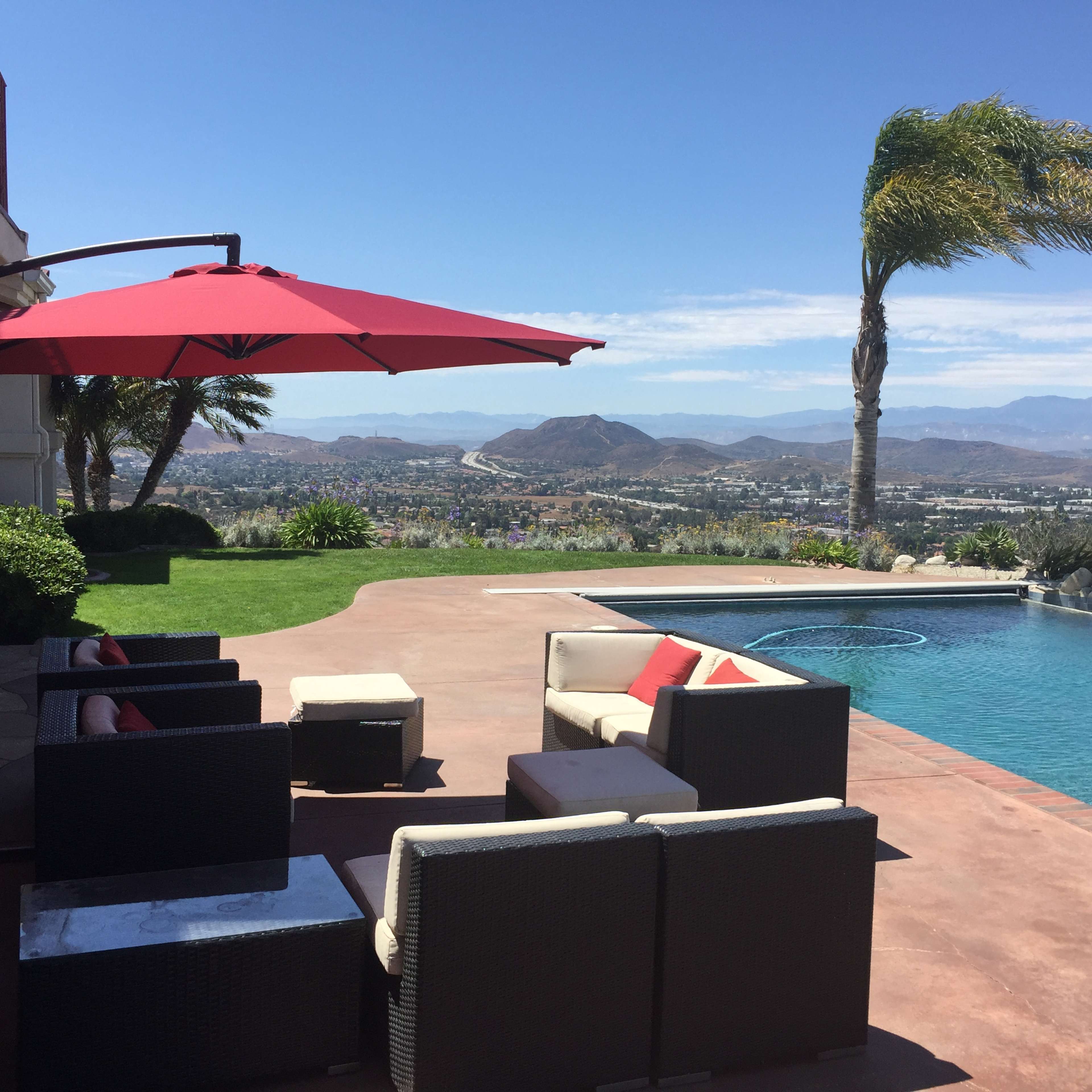 A backyard pool area with lounge chairs and an umbrella, overlooking a scenic view of mountains and a valley.
