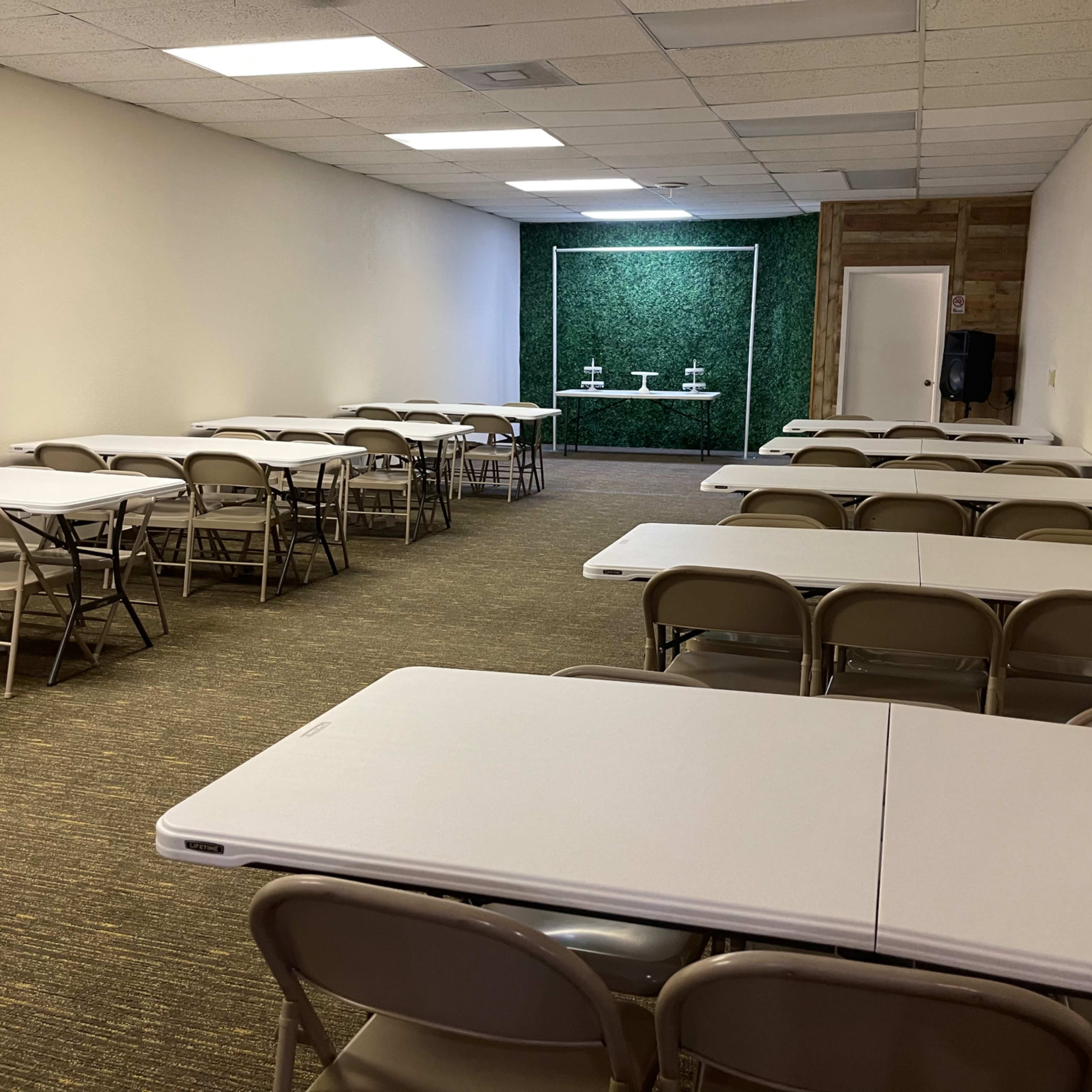 An empty meeting room with multiple folding tables and chairs arranged in rows, a green wall feature in the background, and a door on the right side.