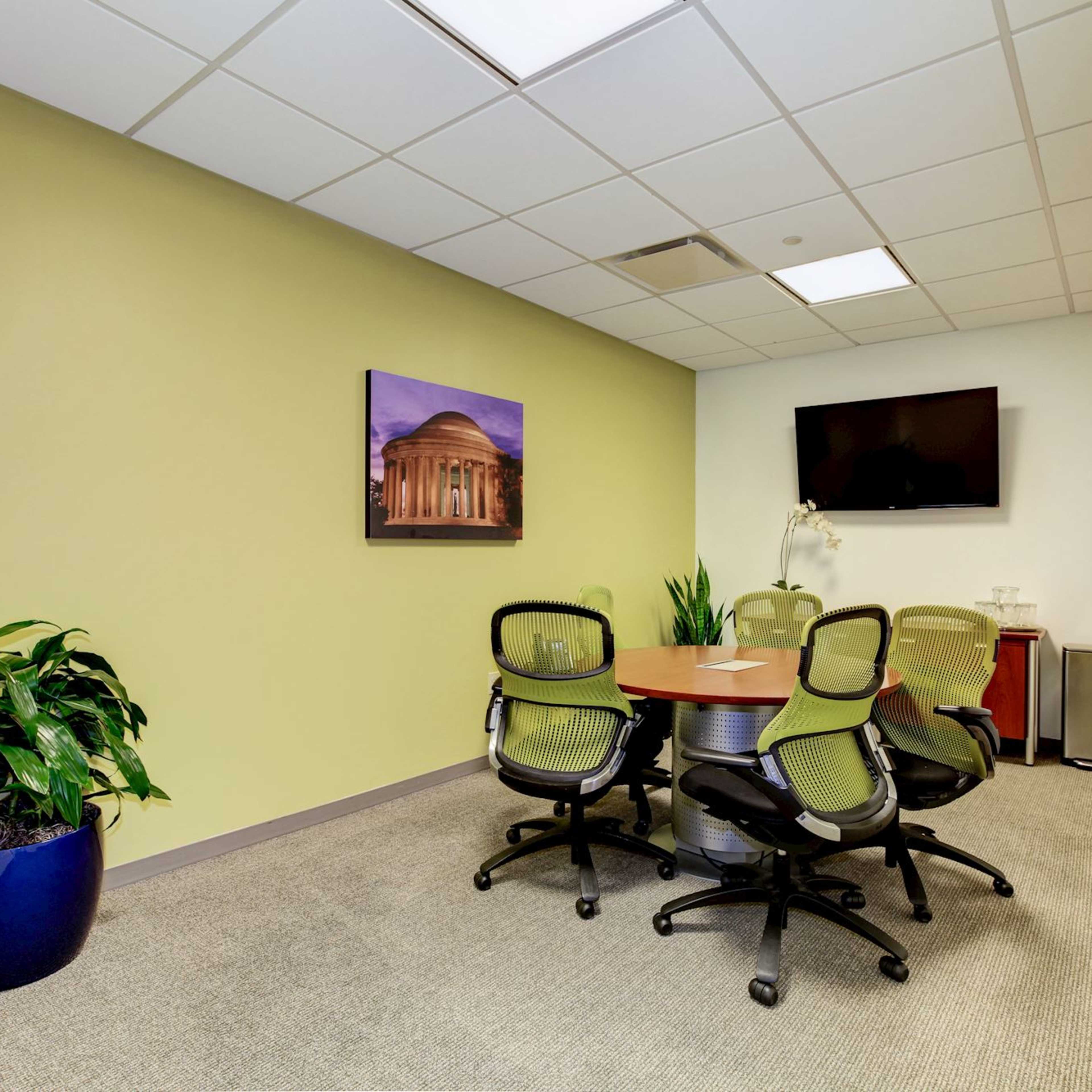 A small meeting room with a round table surrounded by four chairs, a green plant in a blue pot, a flat-screen TV on the wall, and a framed picture of a building.