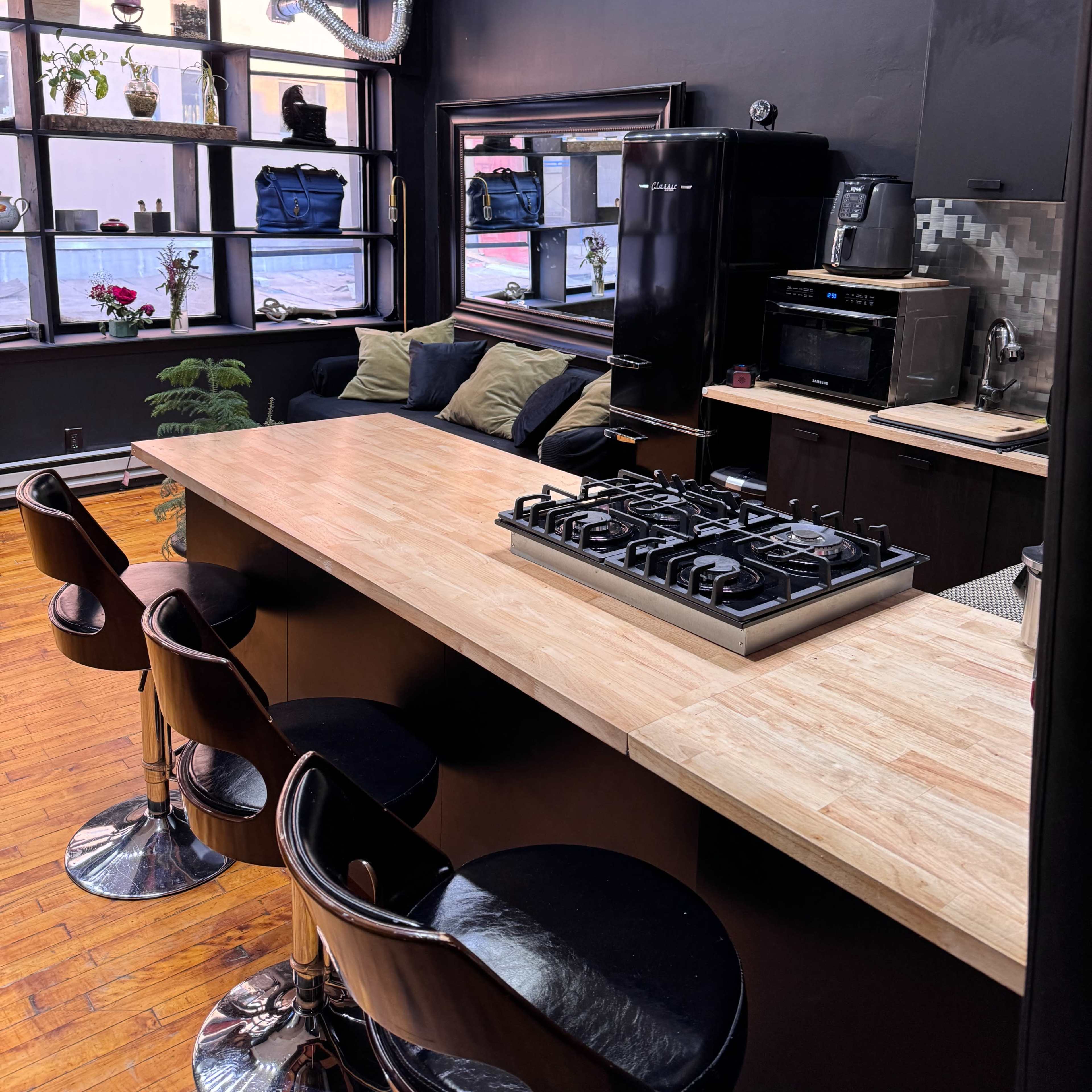A modern kitchen features a central island with a gas stove, bar stools, and black appliances against a dark wall with large windows.