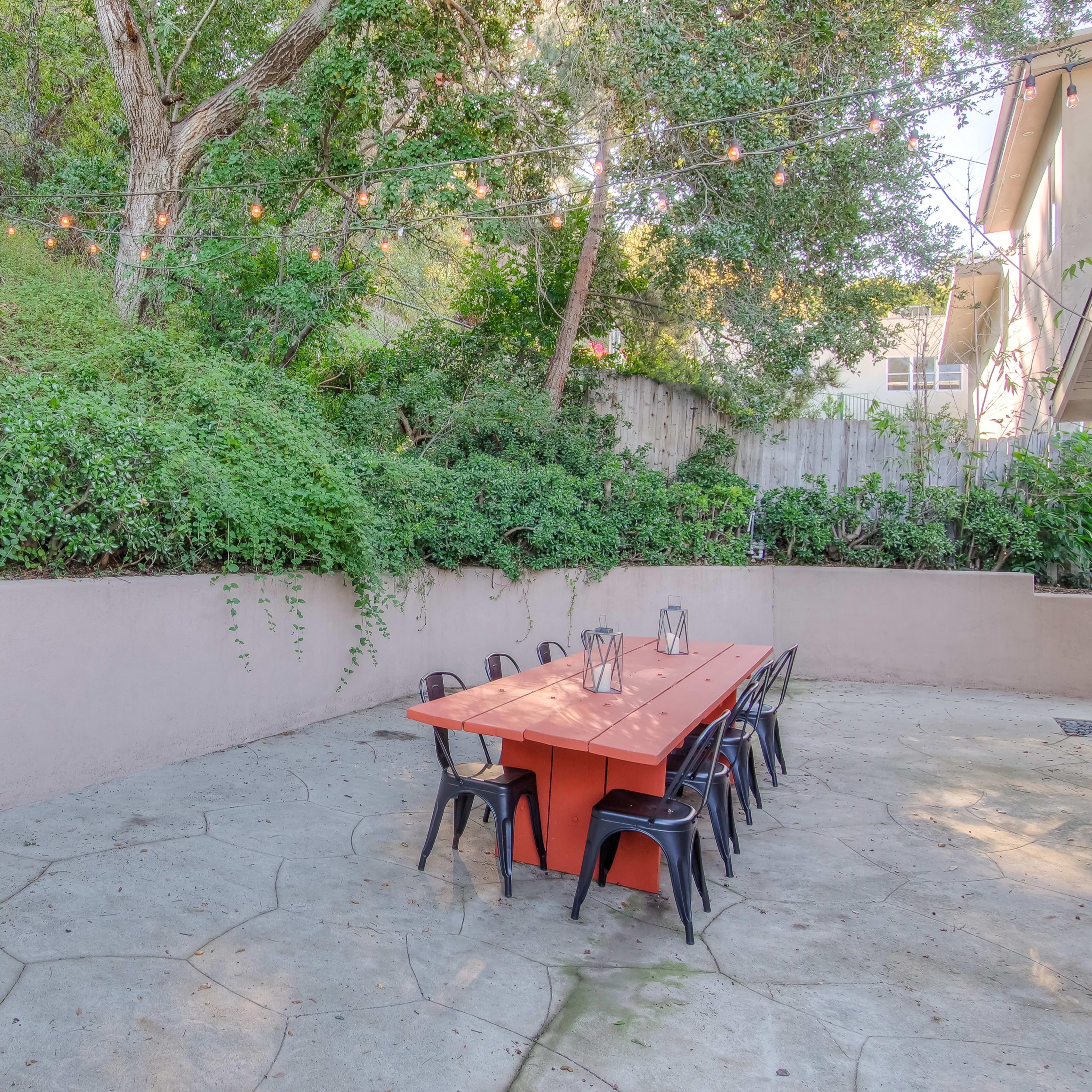 A concrete outdoor dining area with a long red table surrounded by black chairs, set against a backdrop of green foliage and string lights overhead.