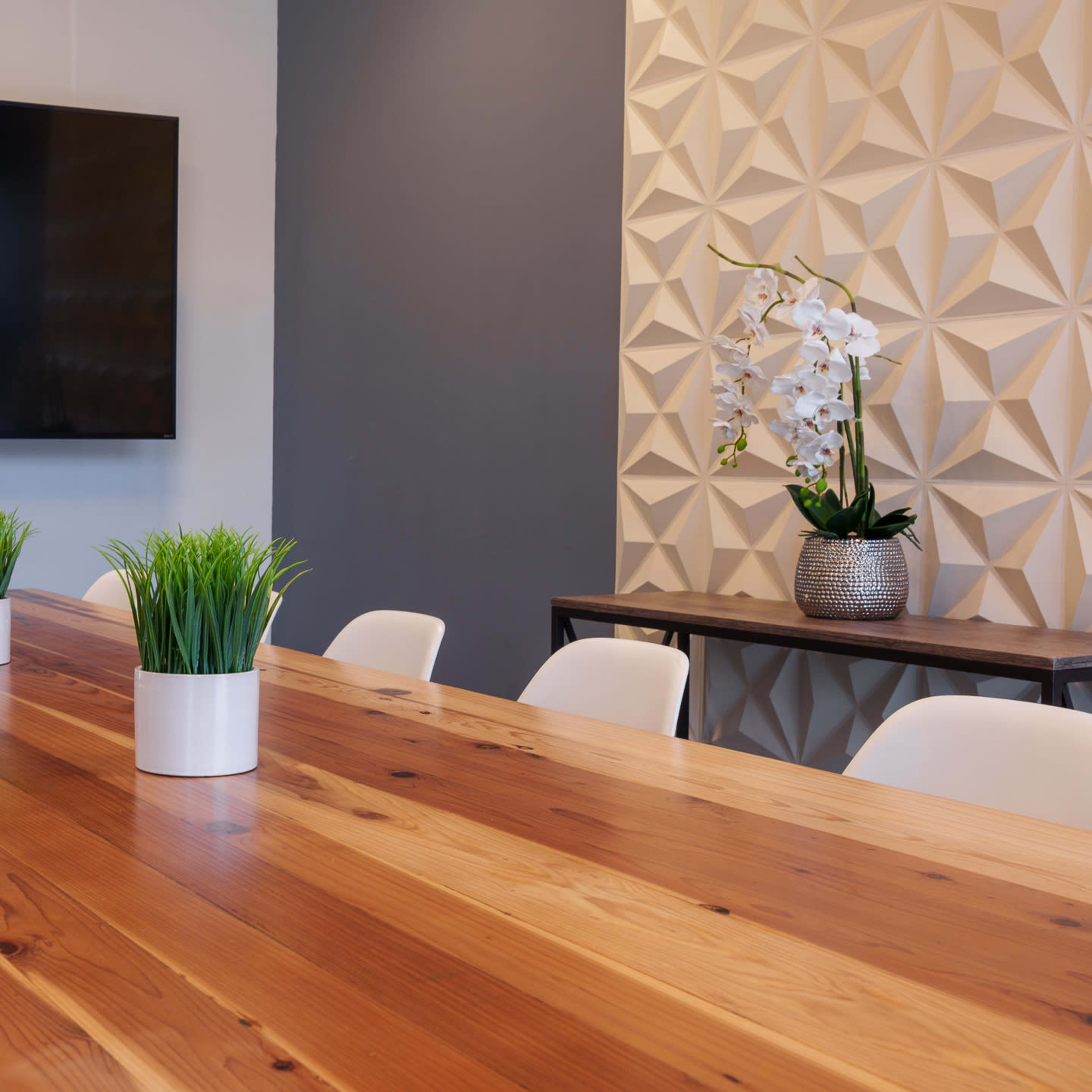 A large wooden conference table with white chairs is set in a meeting room featuring a wall-mounted television and decorative plants.