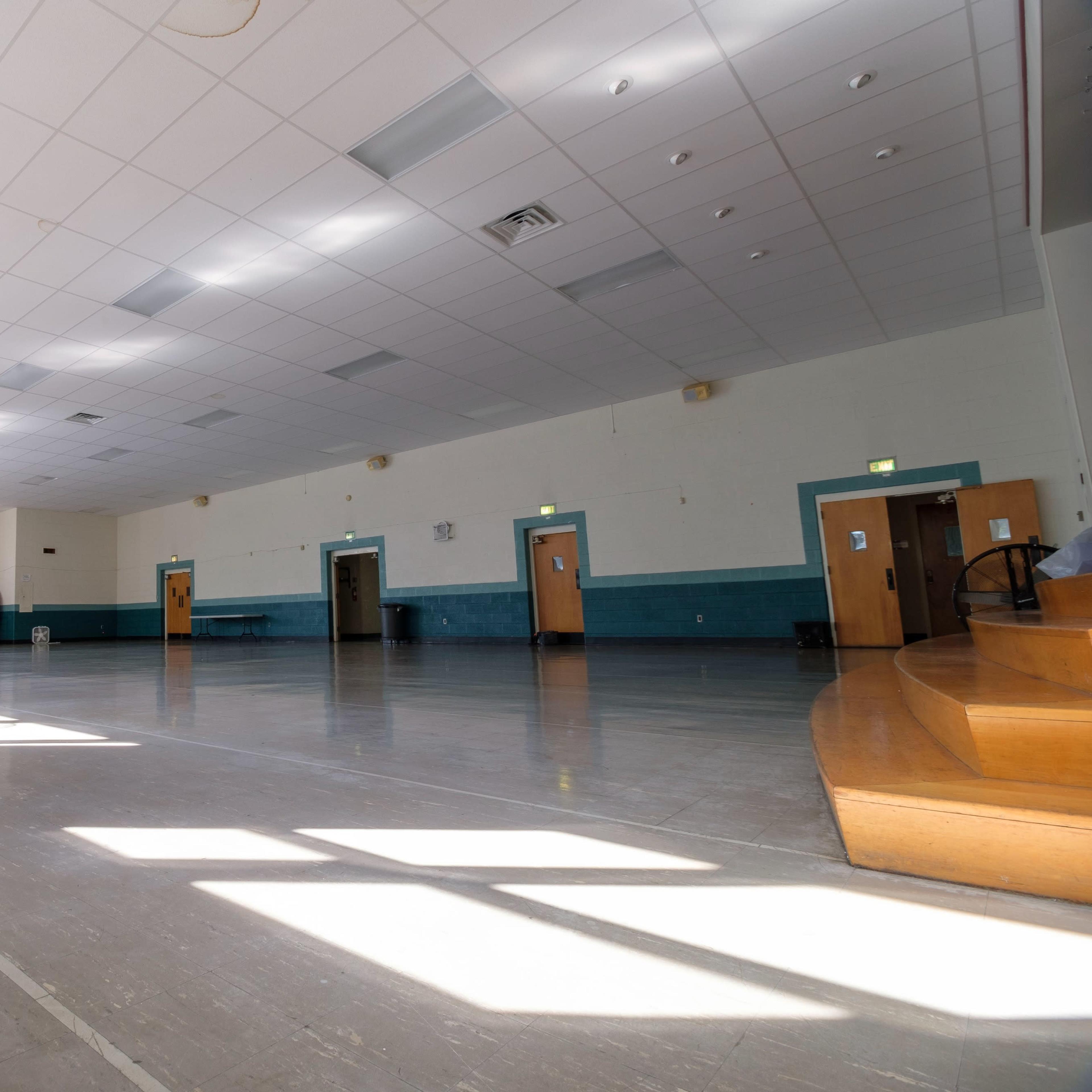 A large, empty indoor space with a shiny floor, multiple doors, and natural light streaming through the windows.