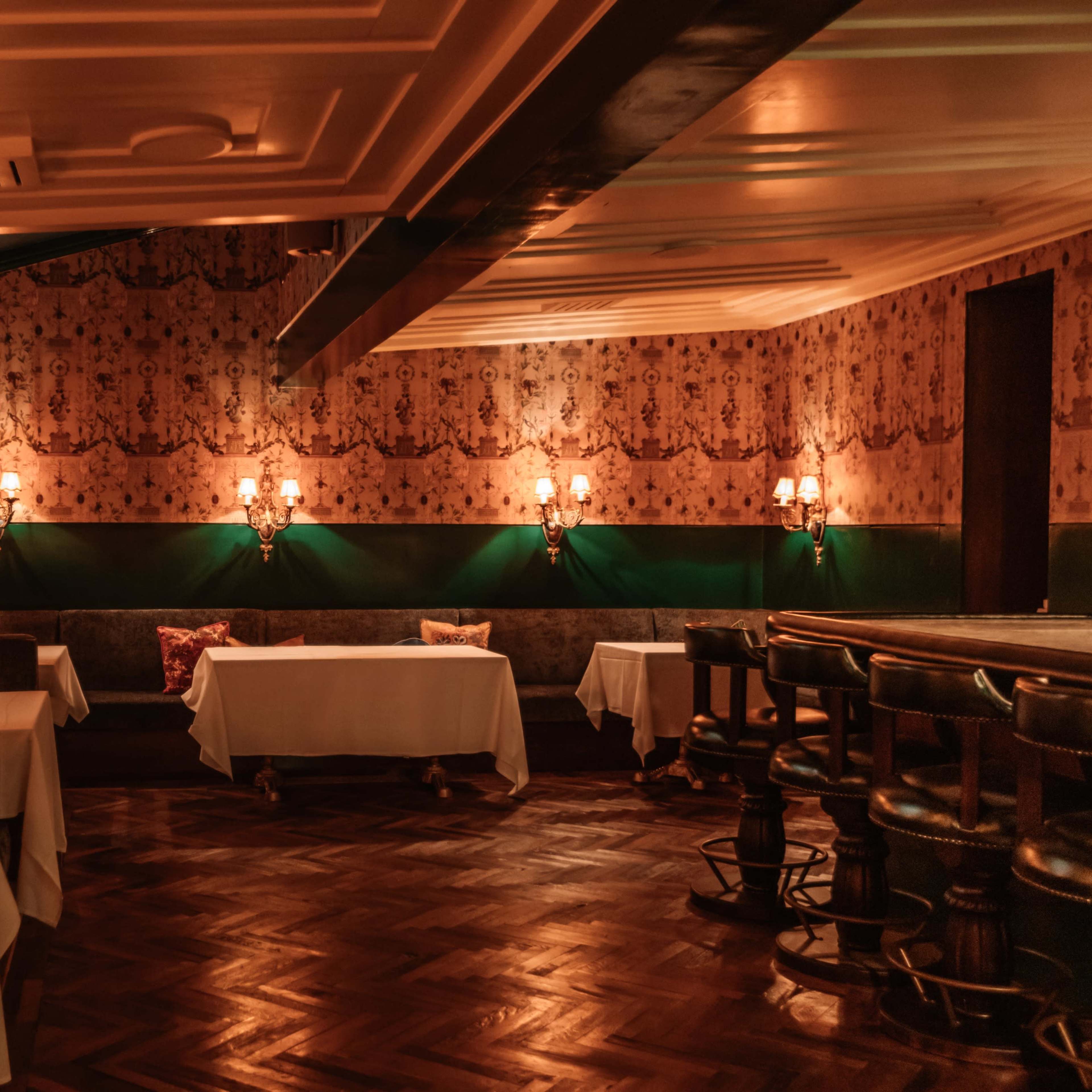 A dimly lit restaurant interior featuring tables with white tablecloths, plush seating along the walls, and elegant wall sconces.