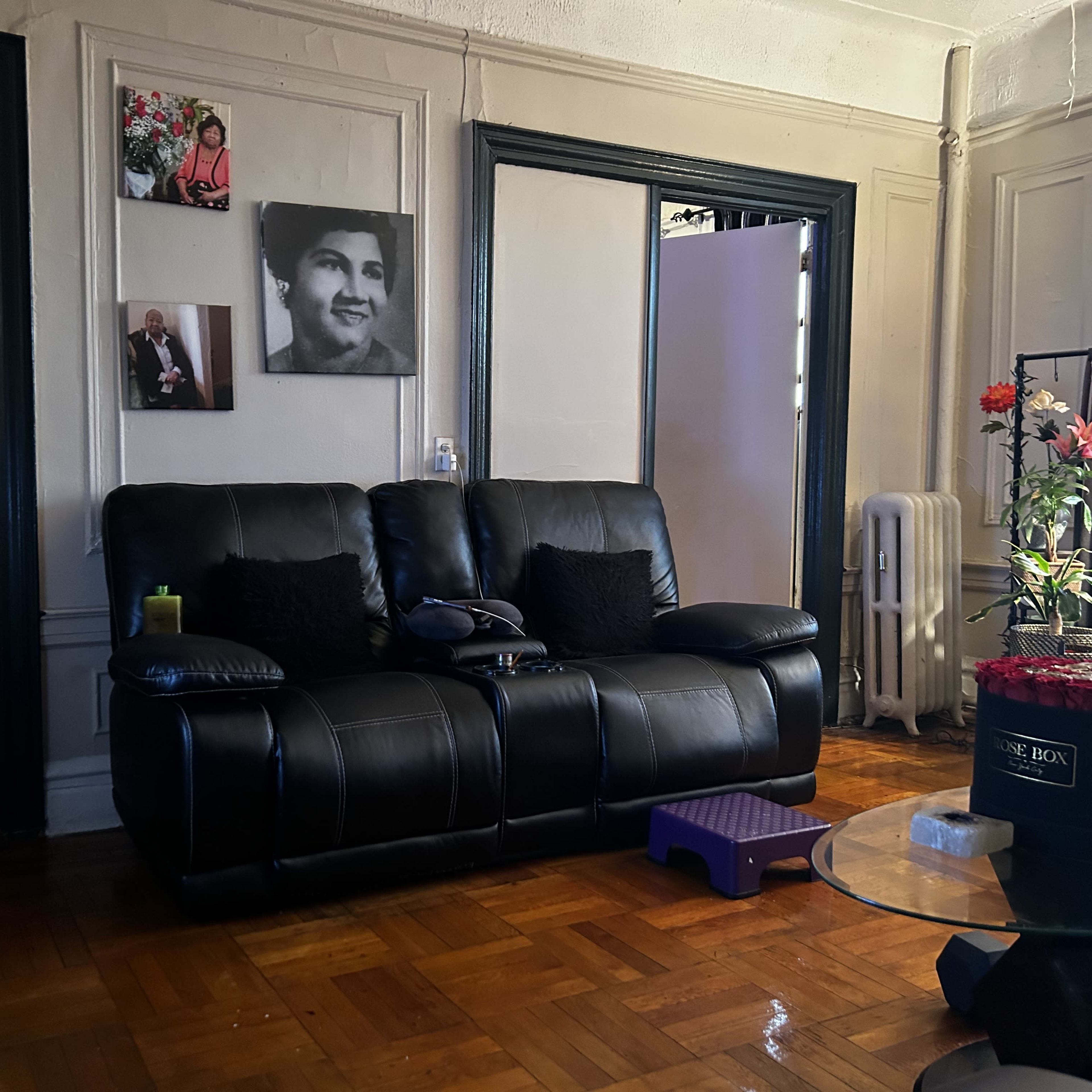 A living room with a black couch, a small purple stool, a glass table, and framed photographs on the wall.