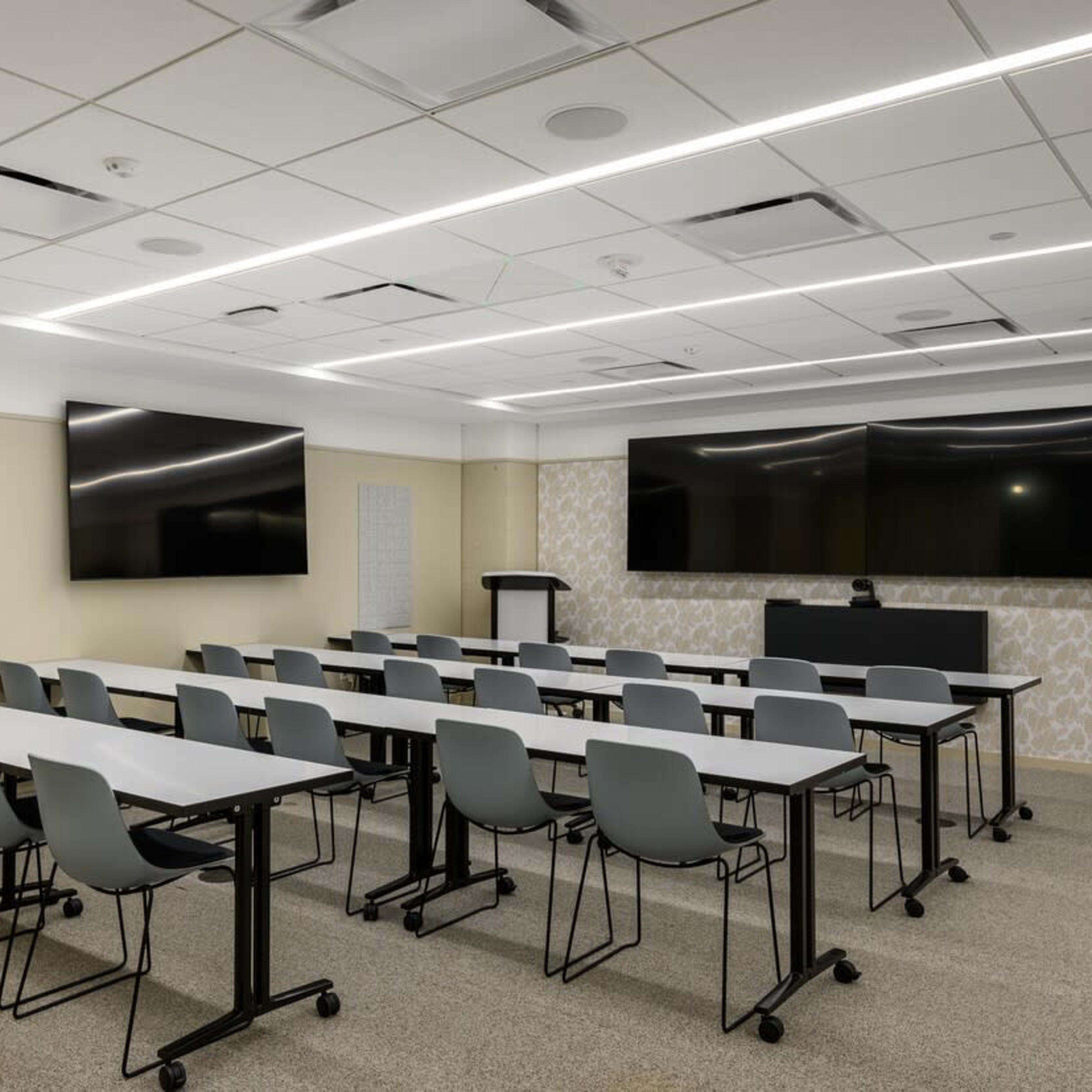 A modern conference room with rows of tables and chairs, two large screens mounted on the wall, and a podium at the front.