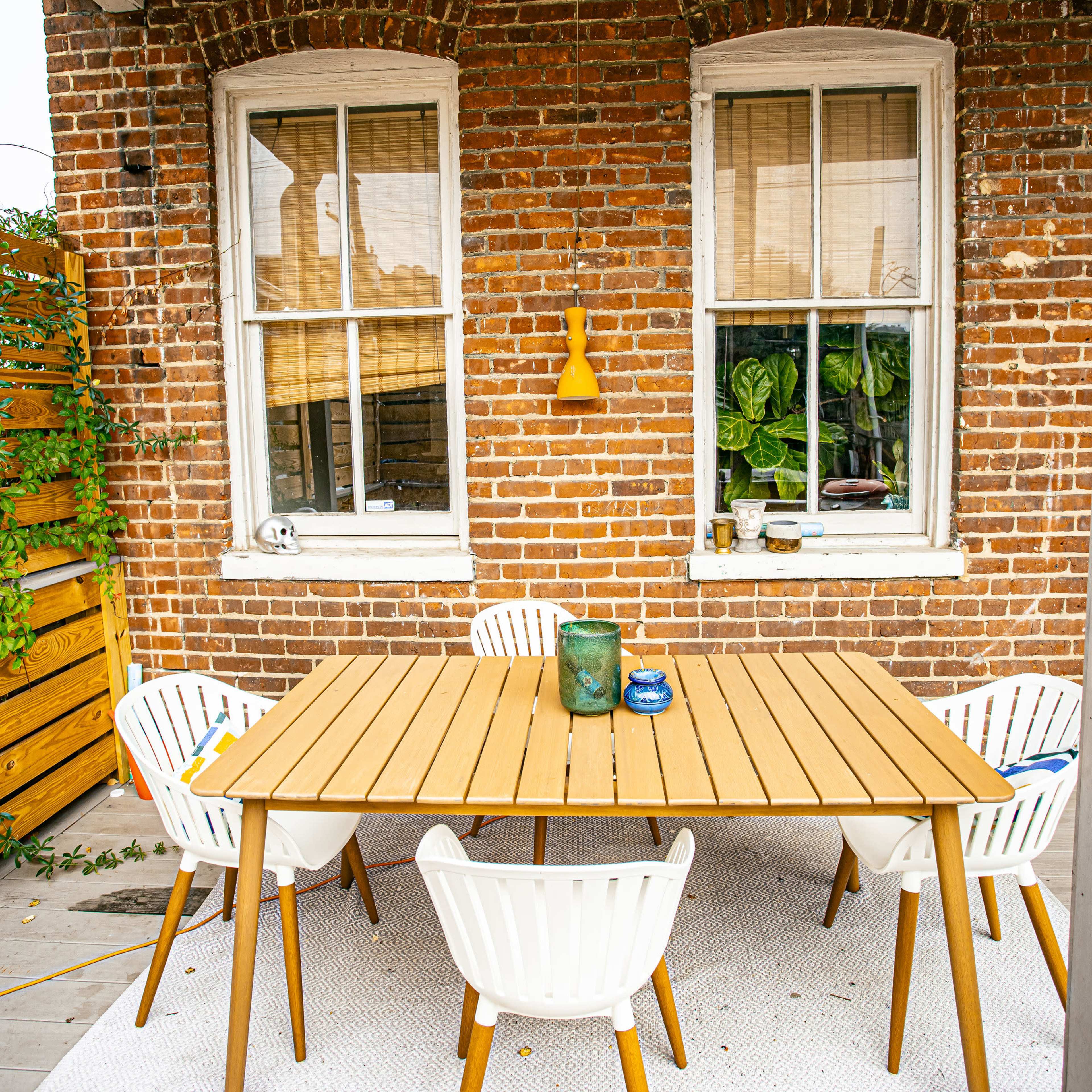 A wooden dining table with four white chairs sits on a light rug beneath a porch with exposed brick walls and two large windows.