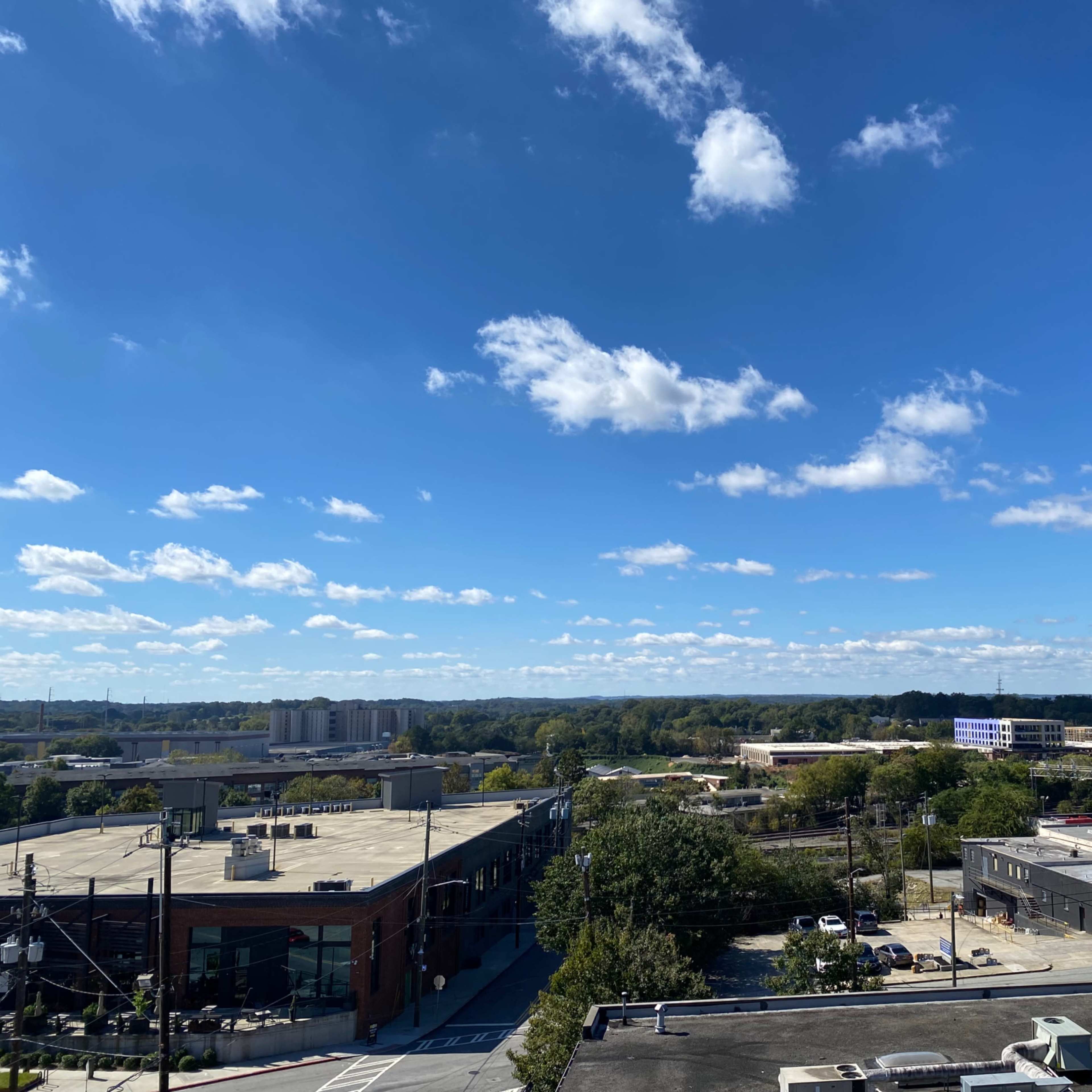 A clear blue sky with scattered white clouds over a suburban area featuring buildings, trees, and roads.