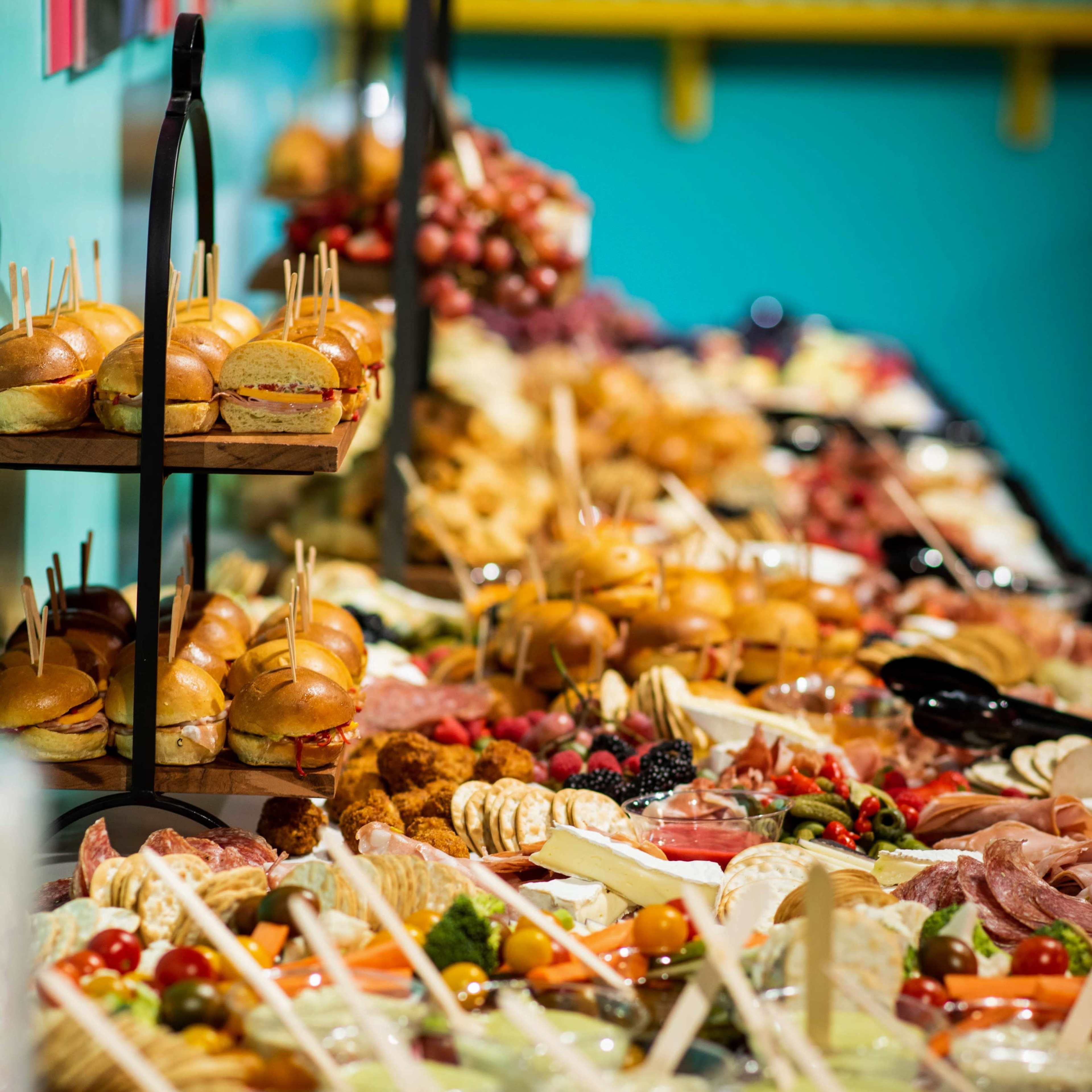 A long table is filled with a variety of food items, including sandwiches, meats, cheeses, fruits, and pastries, arranged for a buffet-style display.