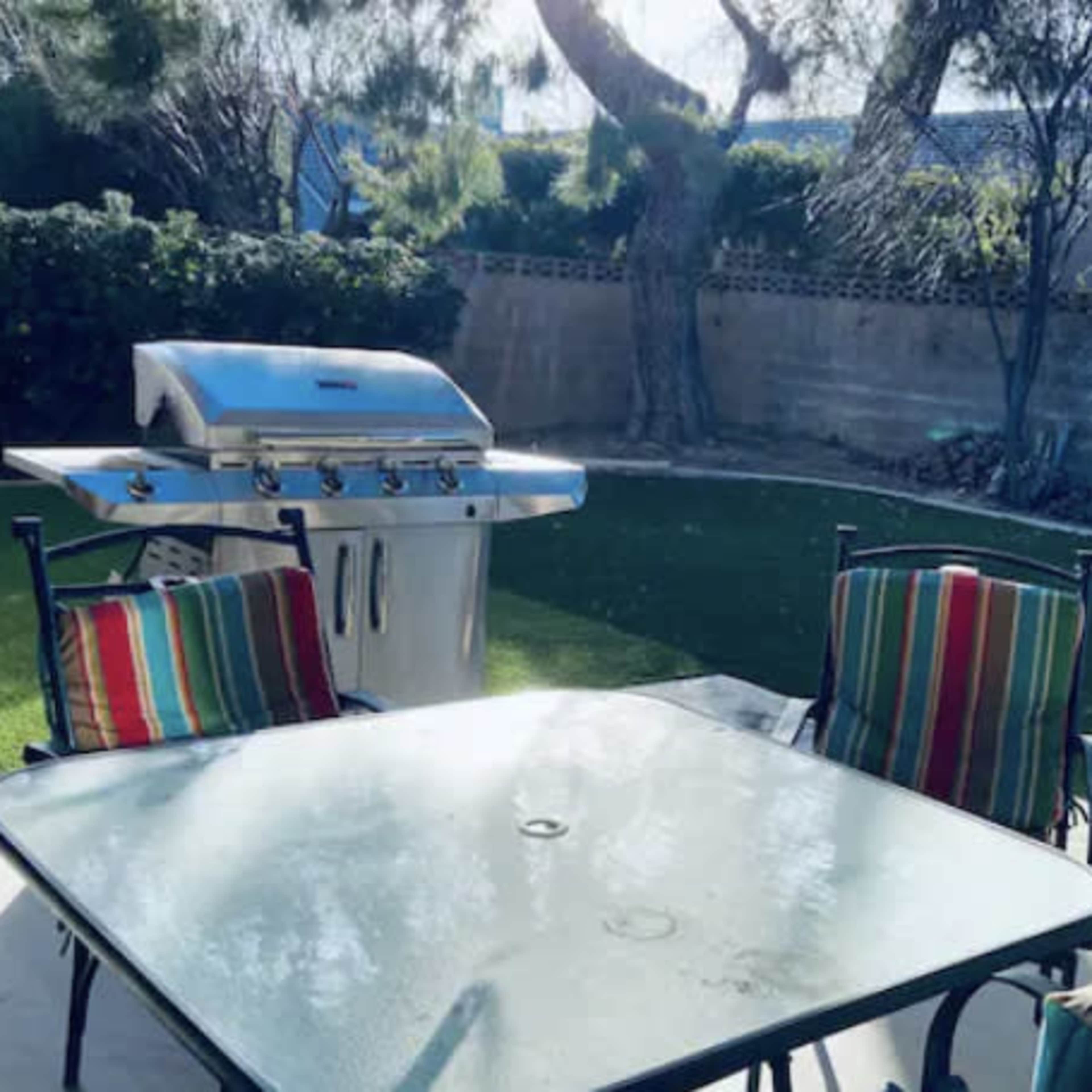 A patio with a glass table and striped chairs overlooks a grassy area and a silver barbecue grill.