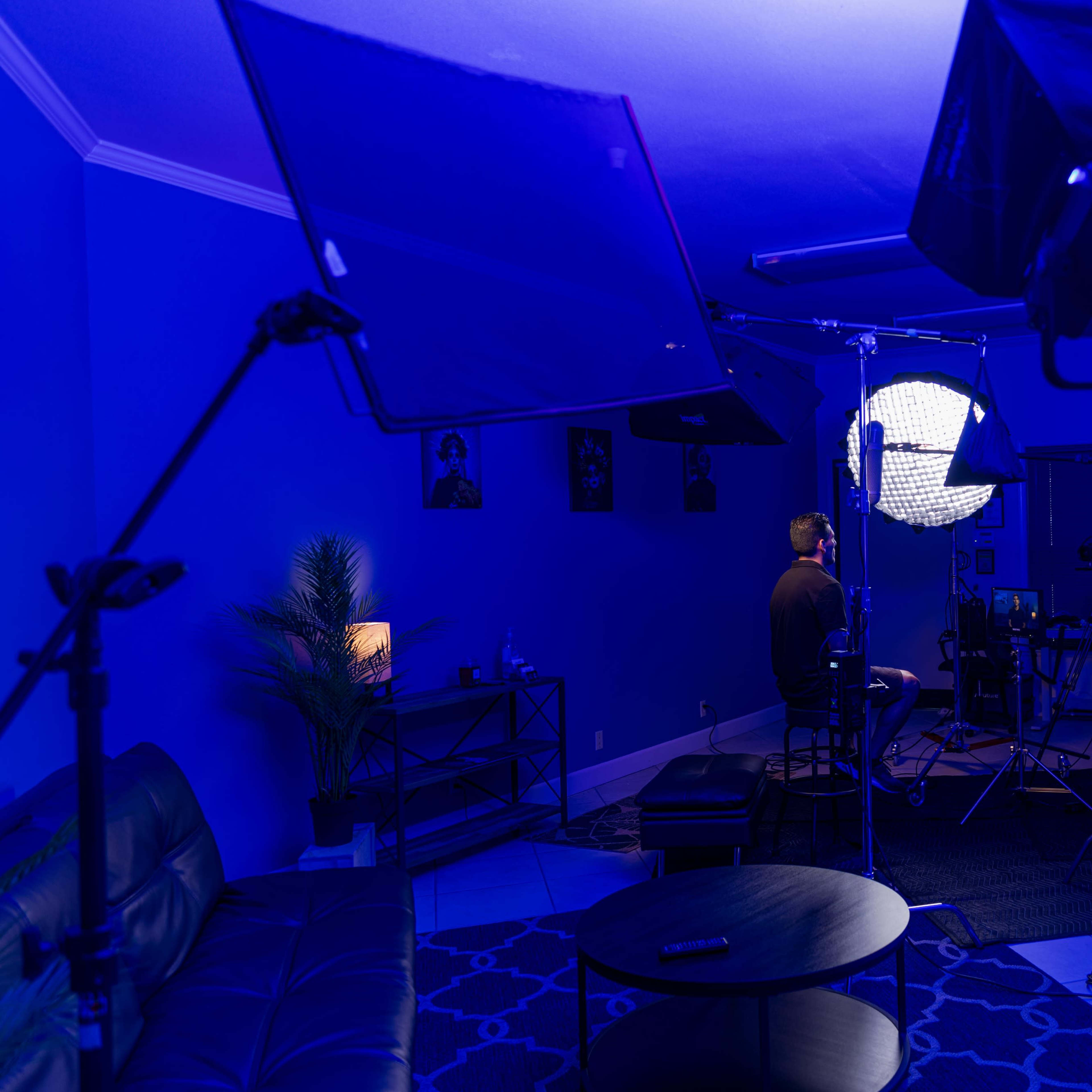 A person sits on a stool in a dimly lit studio space surrounded by photography lights and equipment.