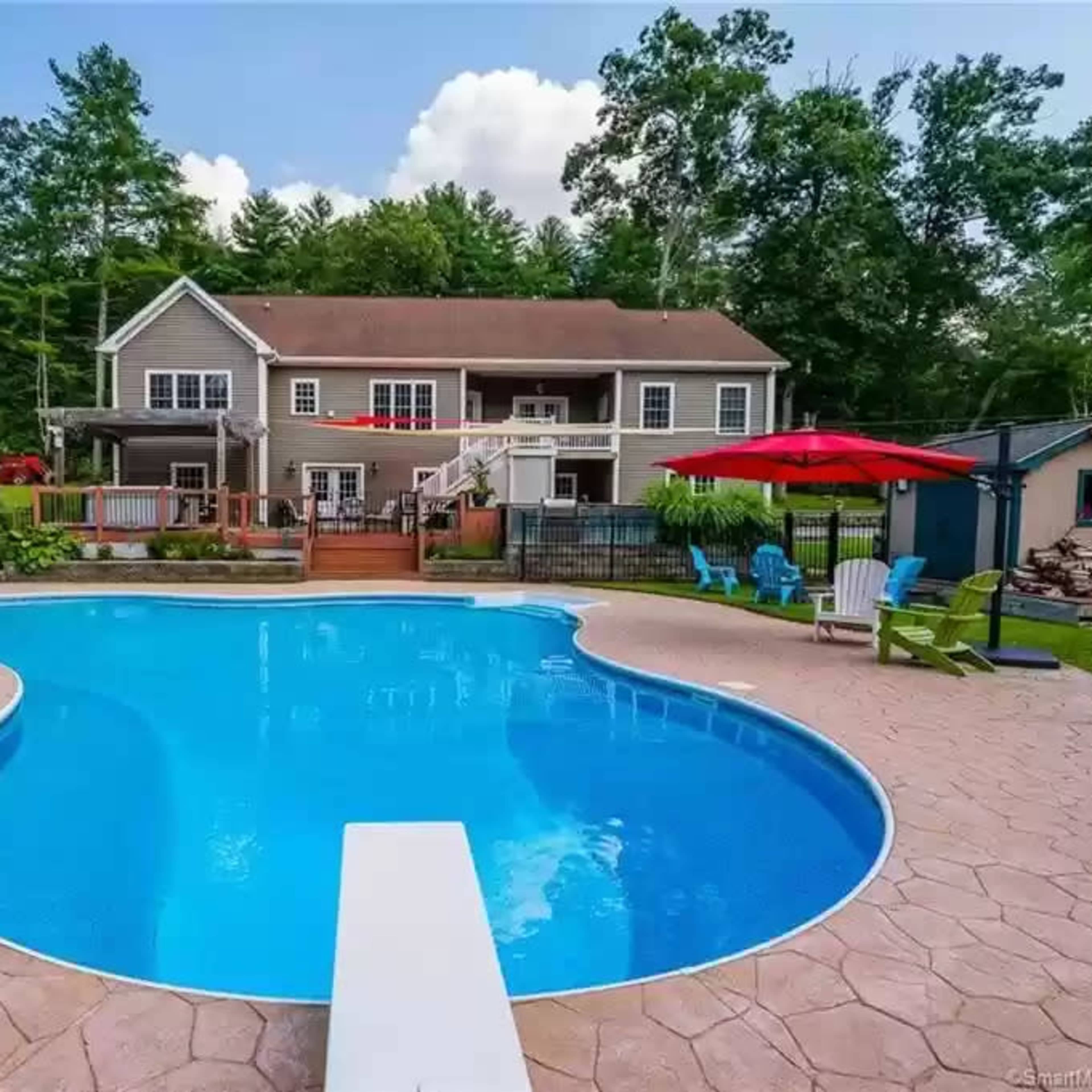 A backyard swimming pool with a diving board, surrounded by a patio and green trees, with a house in the background.