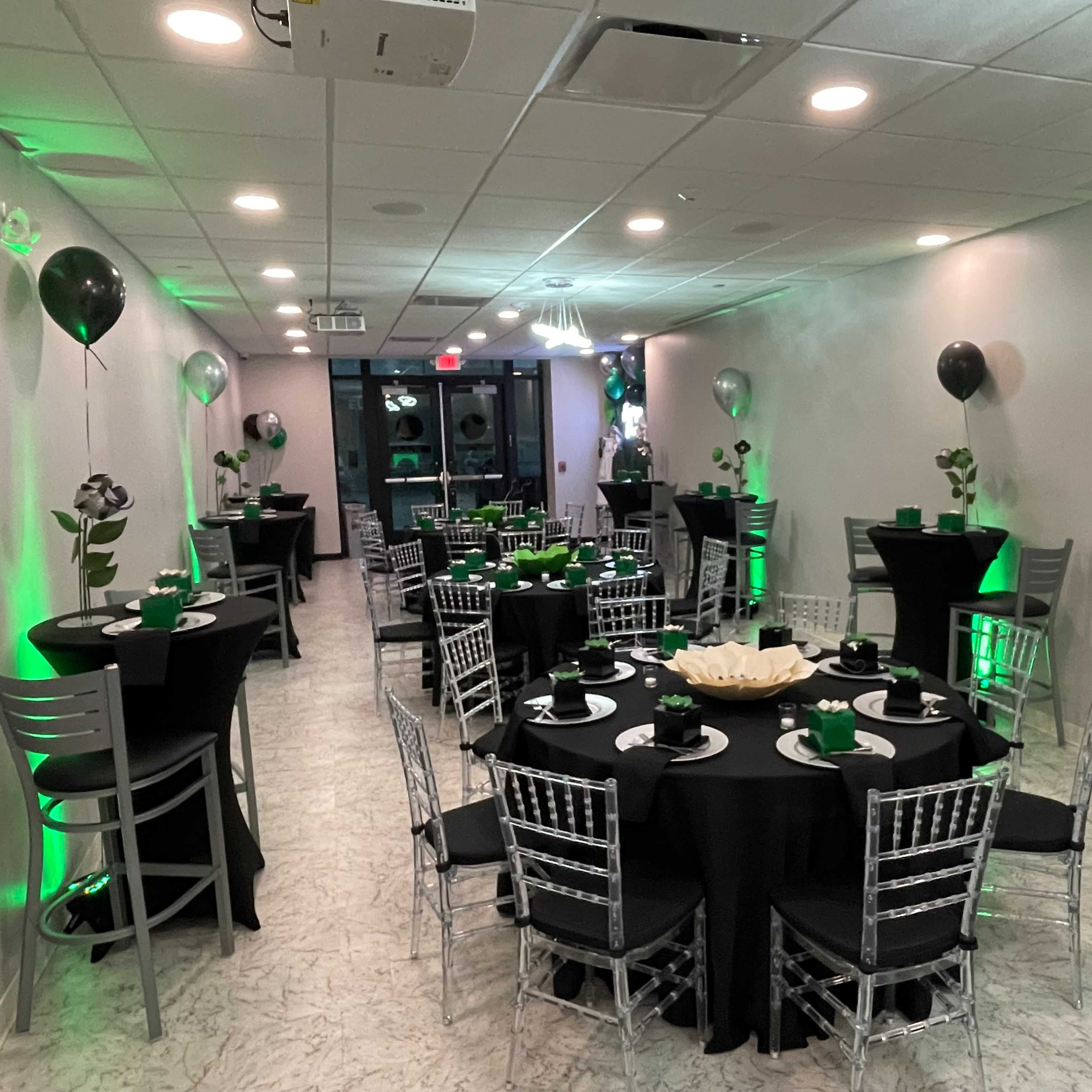 A banquet hall set up for an event with round tables covered in black cloth, decorated with greenery, and surrounded by silver and black balloons.