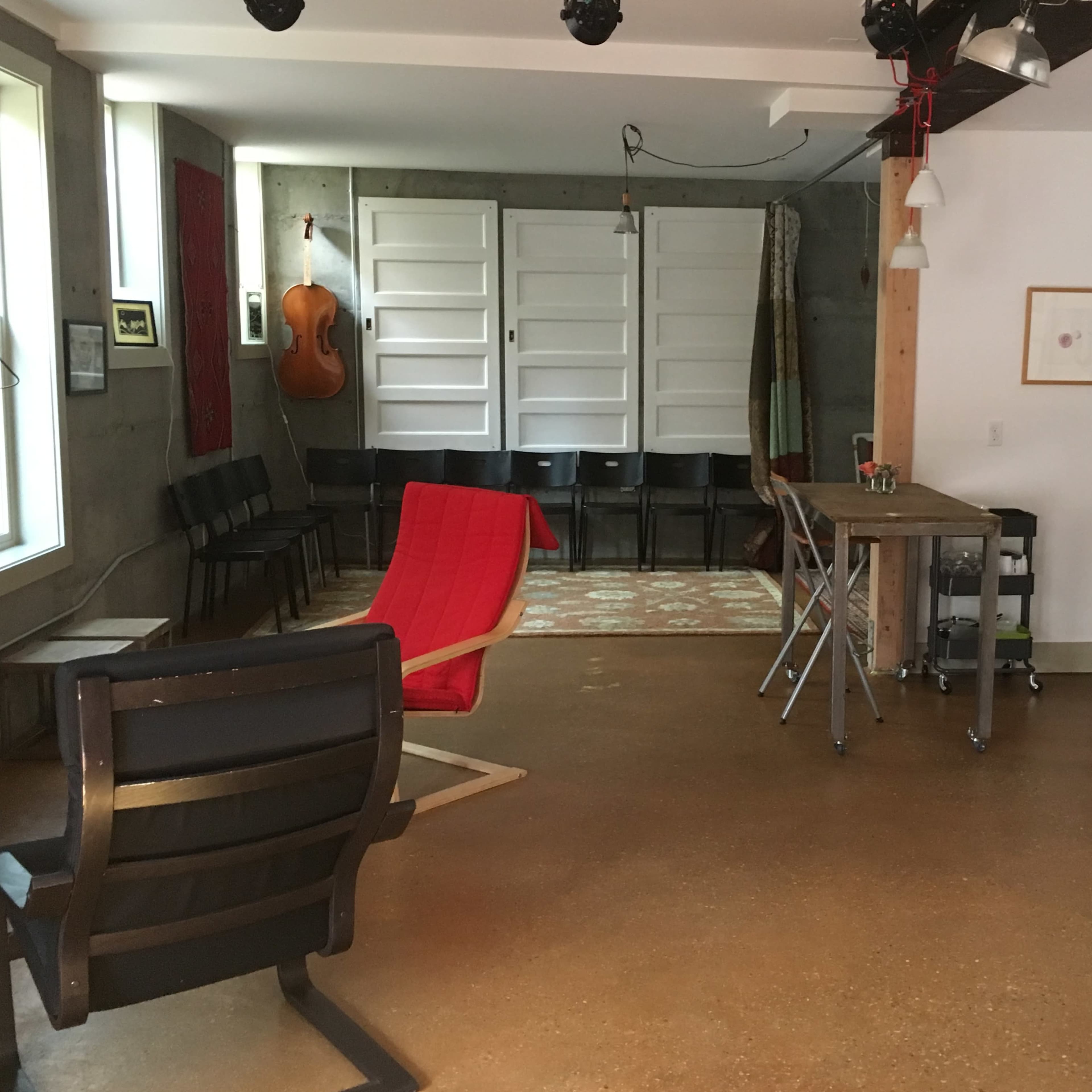 An open interior space featuring a red chair, a wooden table, a kitchenette, and a backdrop of folding doors with a large rug on the floor.