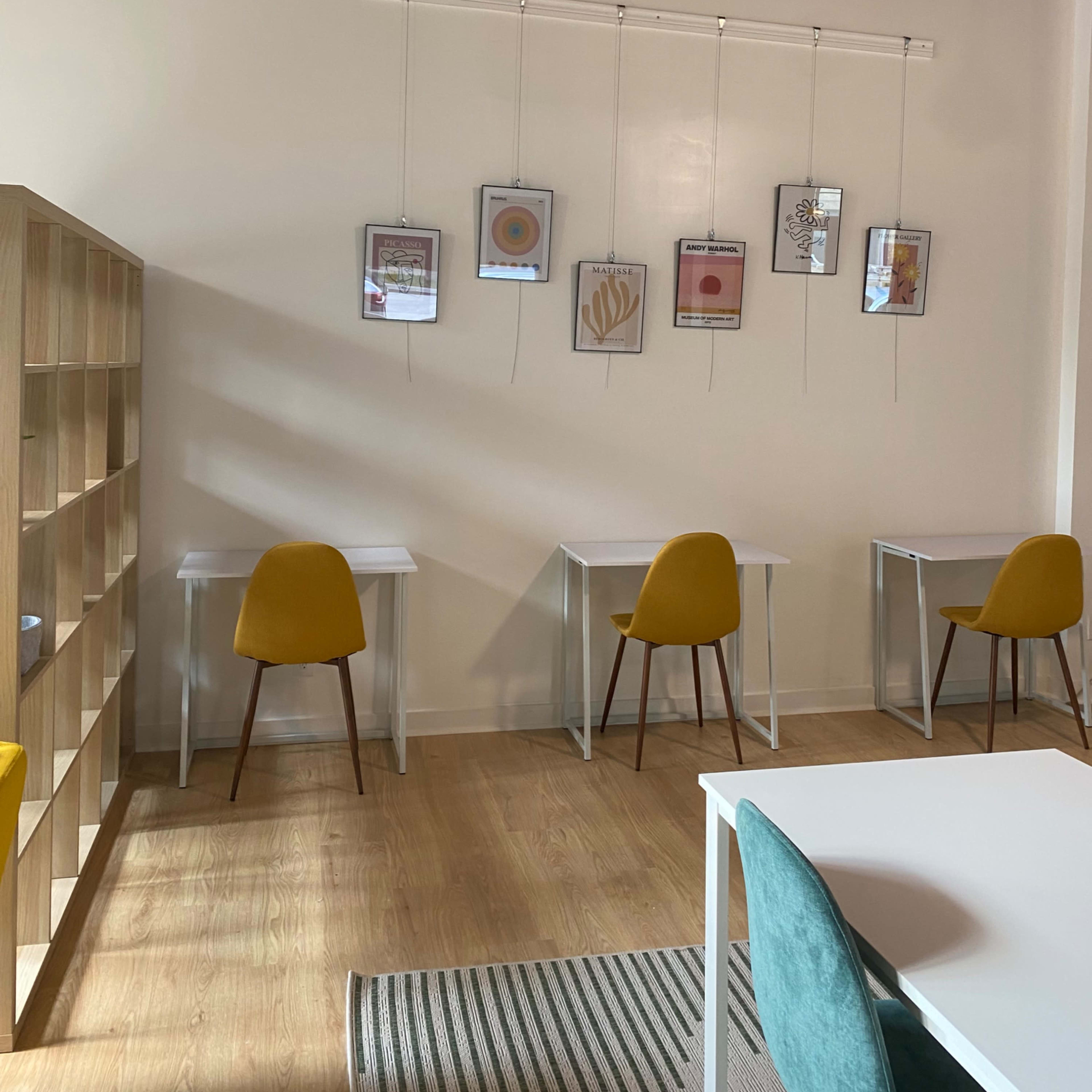 A minimalist workspace with four yellow chairs arranged around white tables, accompanied by framed artworks hanging on the wall above.