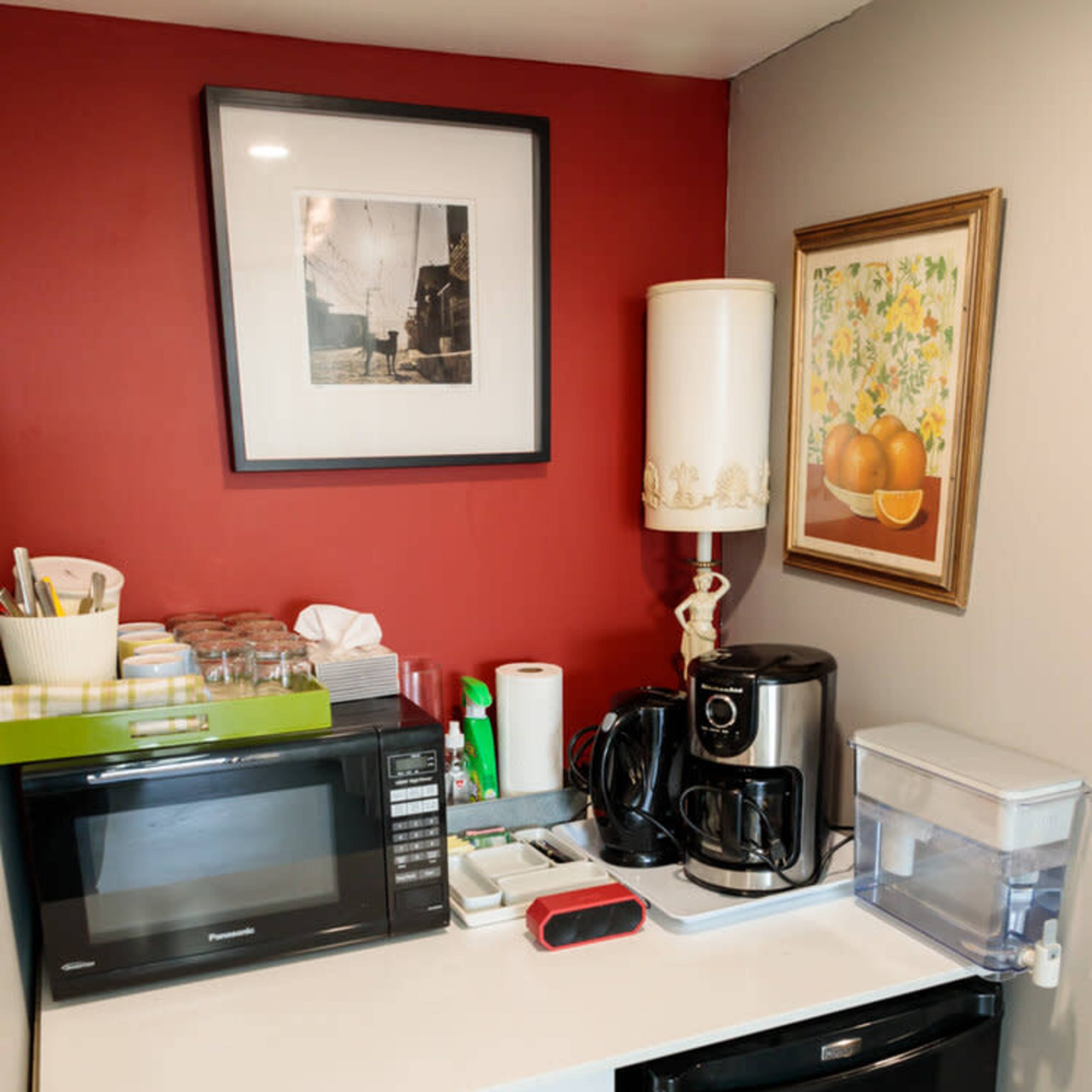 A small kitchen corner featuring a microwave, coffee maker, and a mini fridge, arranged beneath artwork and decorative items.