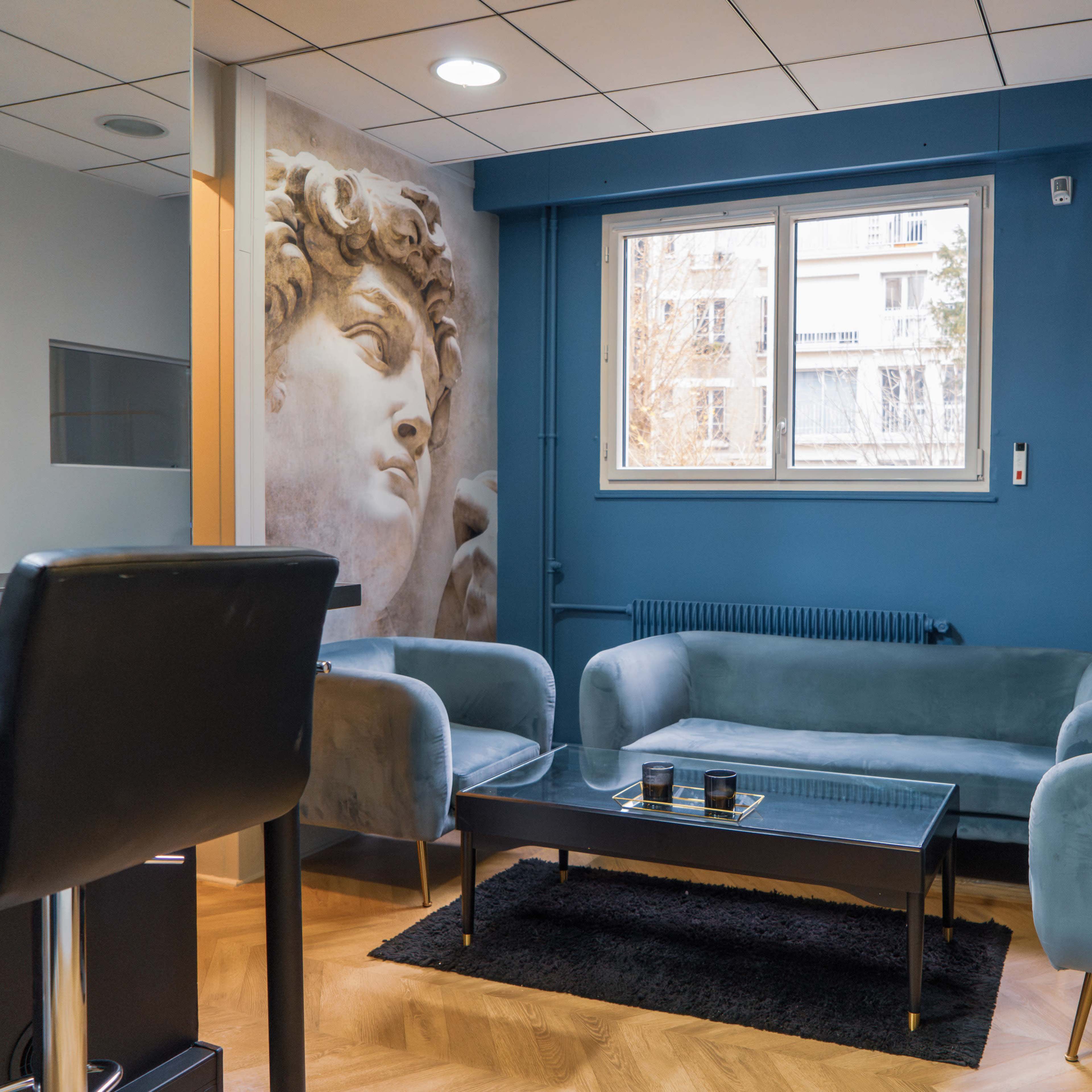 A modern sitting area with two light blue sofas, a black coffee table, and a large mural of a classical sculpture on the wall.