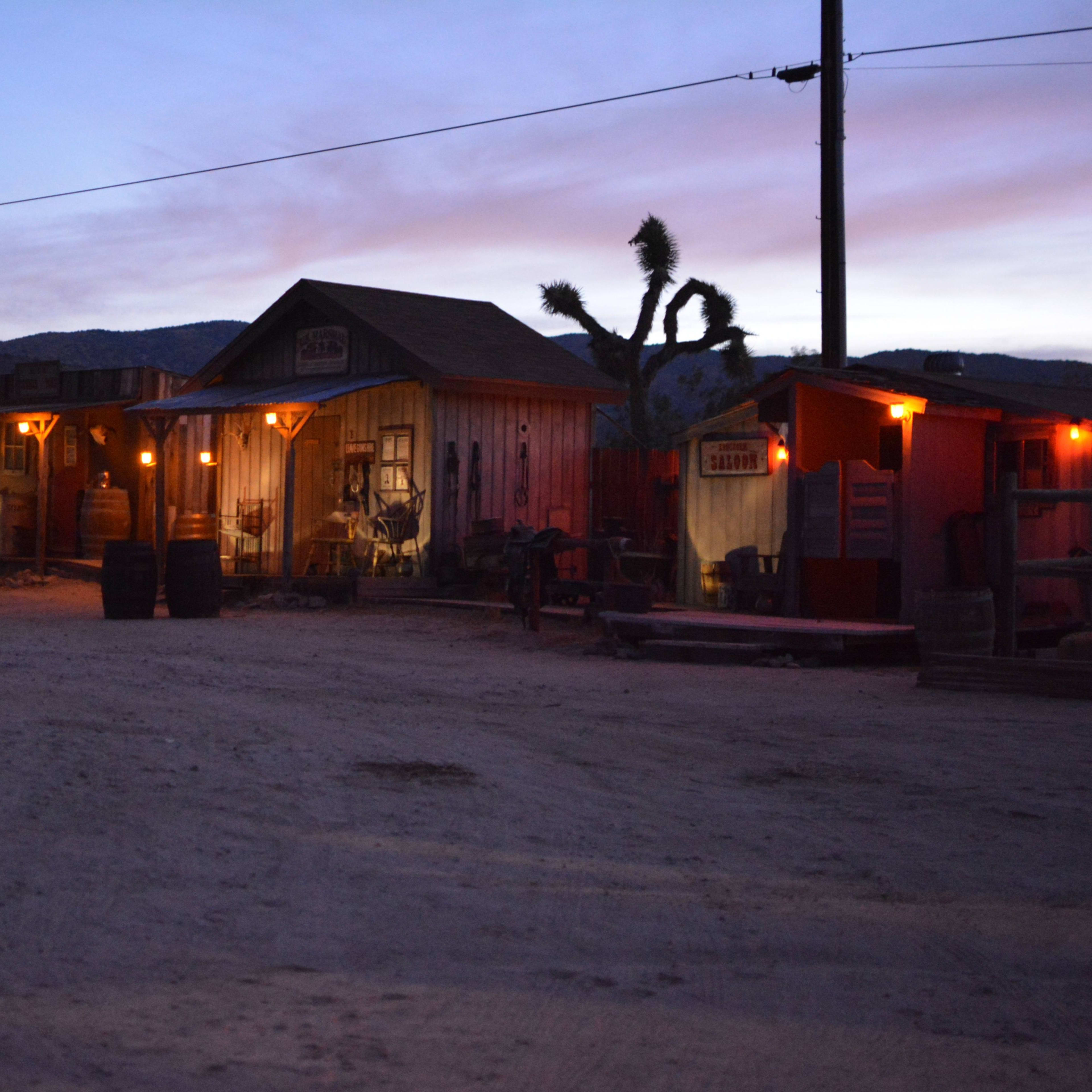 The scene depicts a western-style ghost town at dusk, with illuminated storefronts and a dirt pathway under a colorful sky.