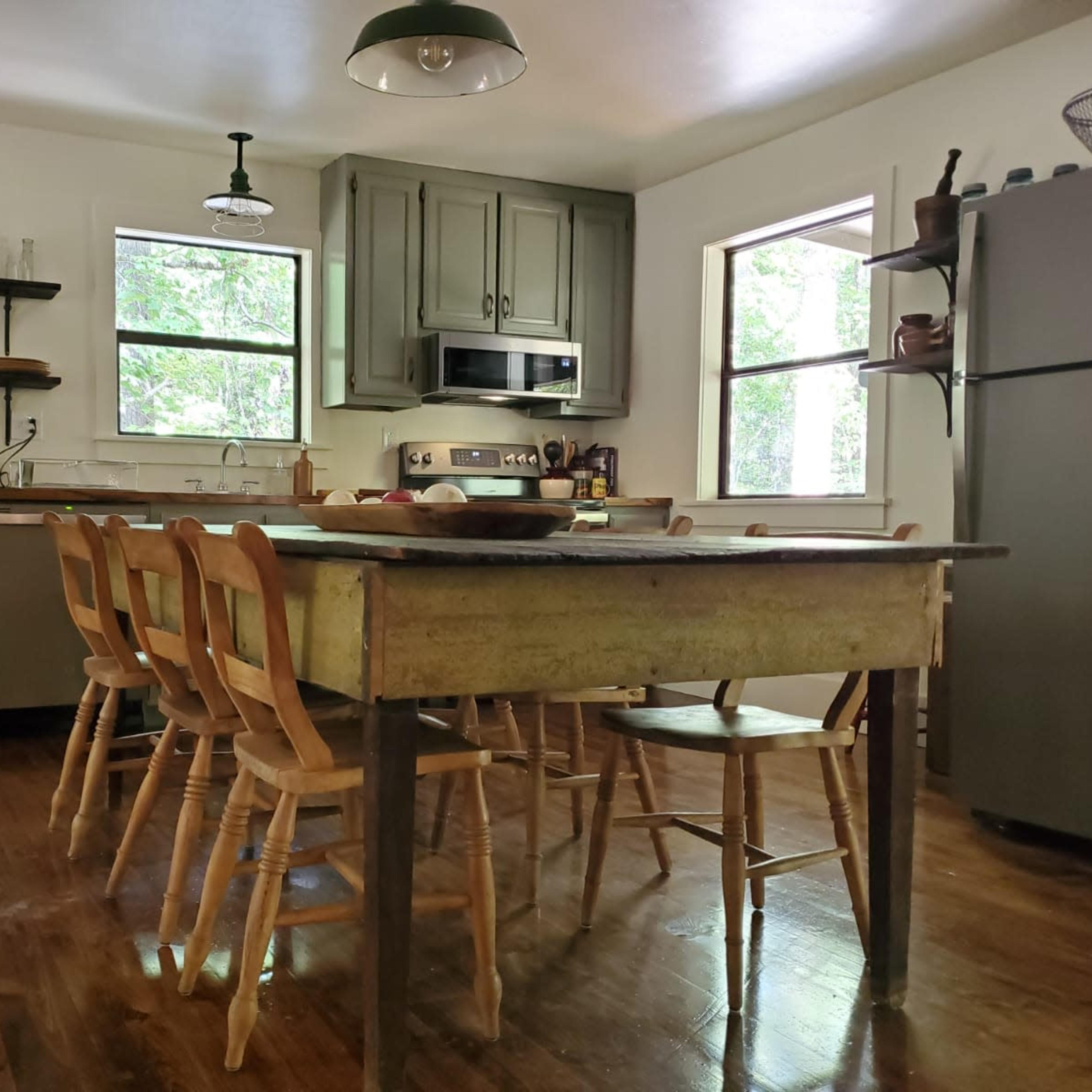 The kitchen features a wooden table surrounded by six chairs, with cabinets, a stove, and shelves displaying dishes against a backdrop of large windows.