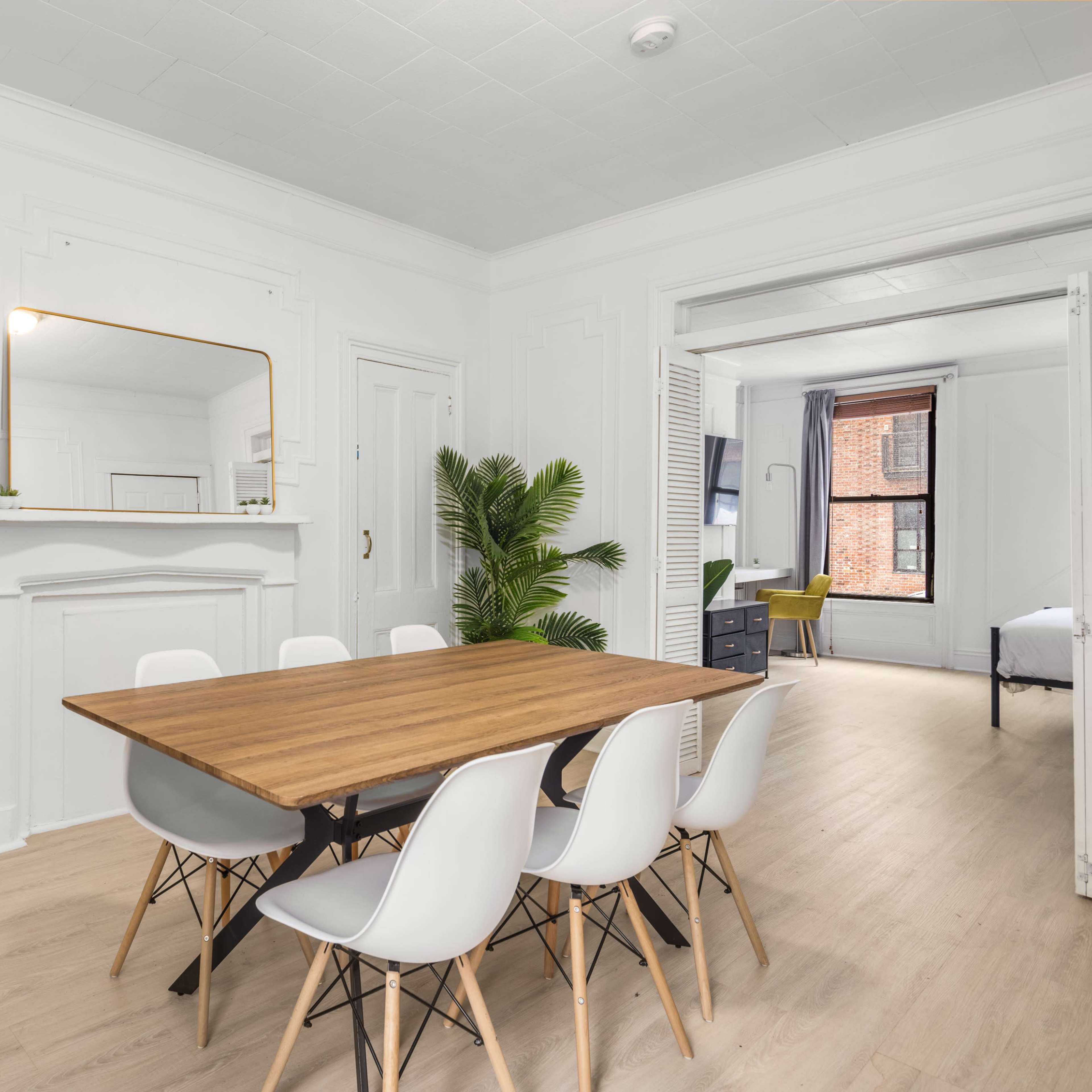A bright dining area with a wooden table surrounded by white chairs, a green plant in a pot, and a gold-framed mirror on the wall.