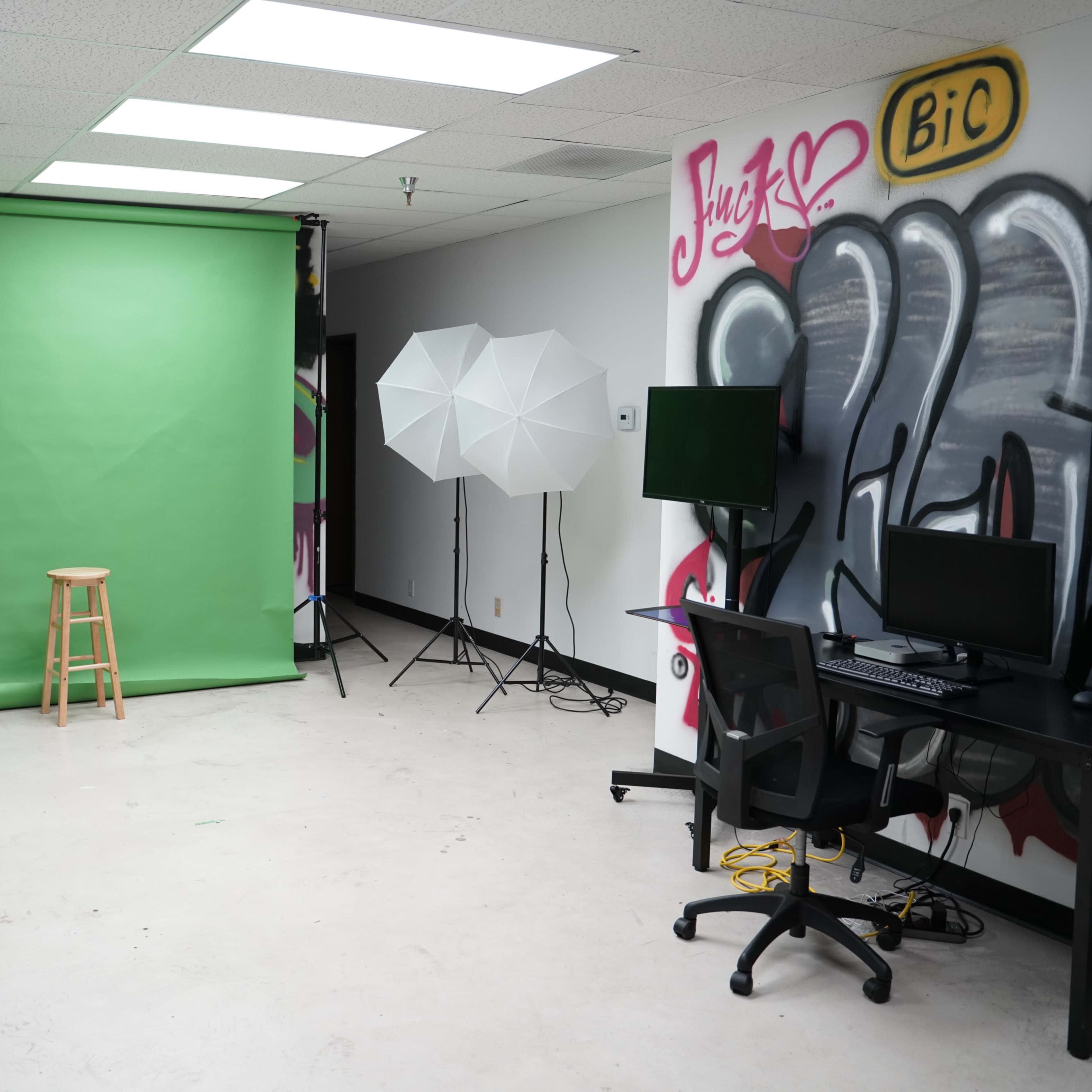 A photo studio featuring a green screen, two light setups, a stool, and a computer workstation against a graffiti-decorated wall.