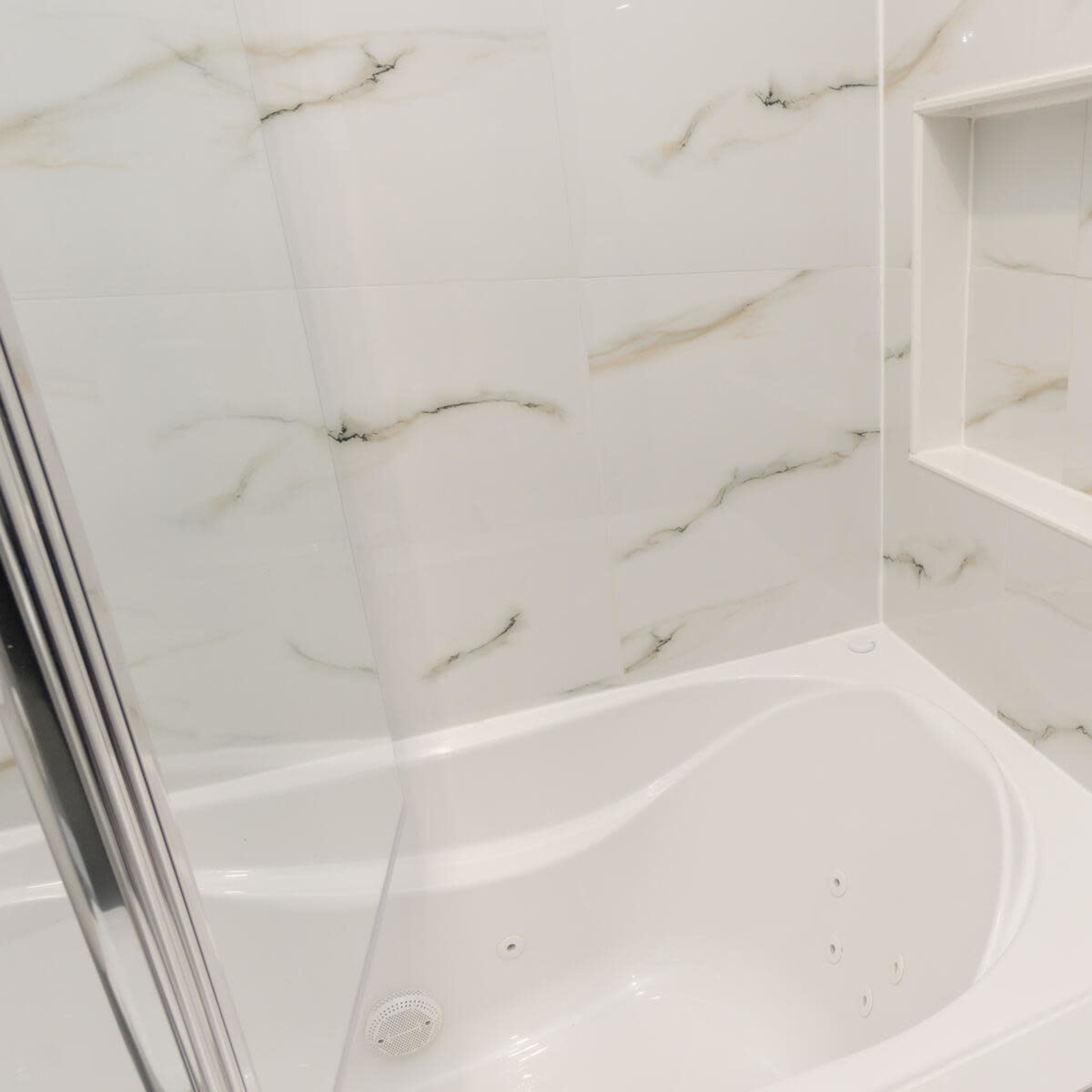 A modern bathroom featuring a white bathtub with a marble-patterned tiled wall and a niche for toiletries.
