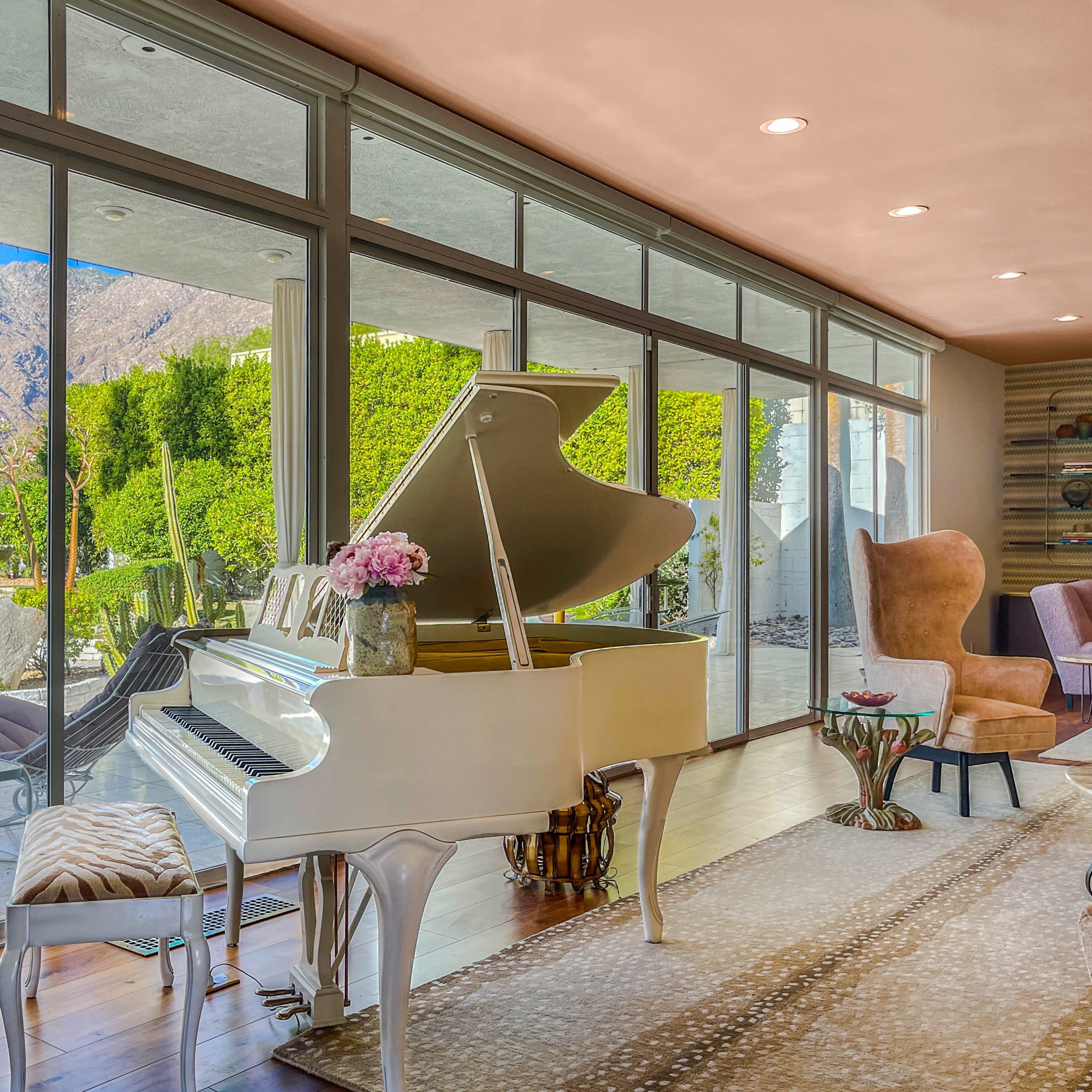 A white grand piano sits beside large windows that overlook a garden and mountains.