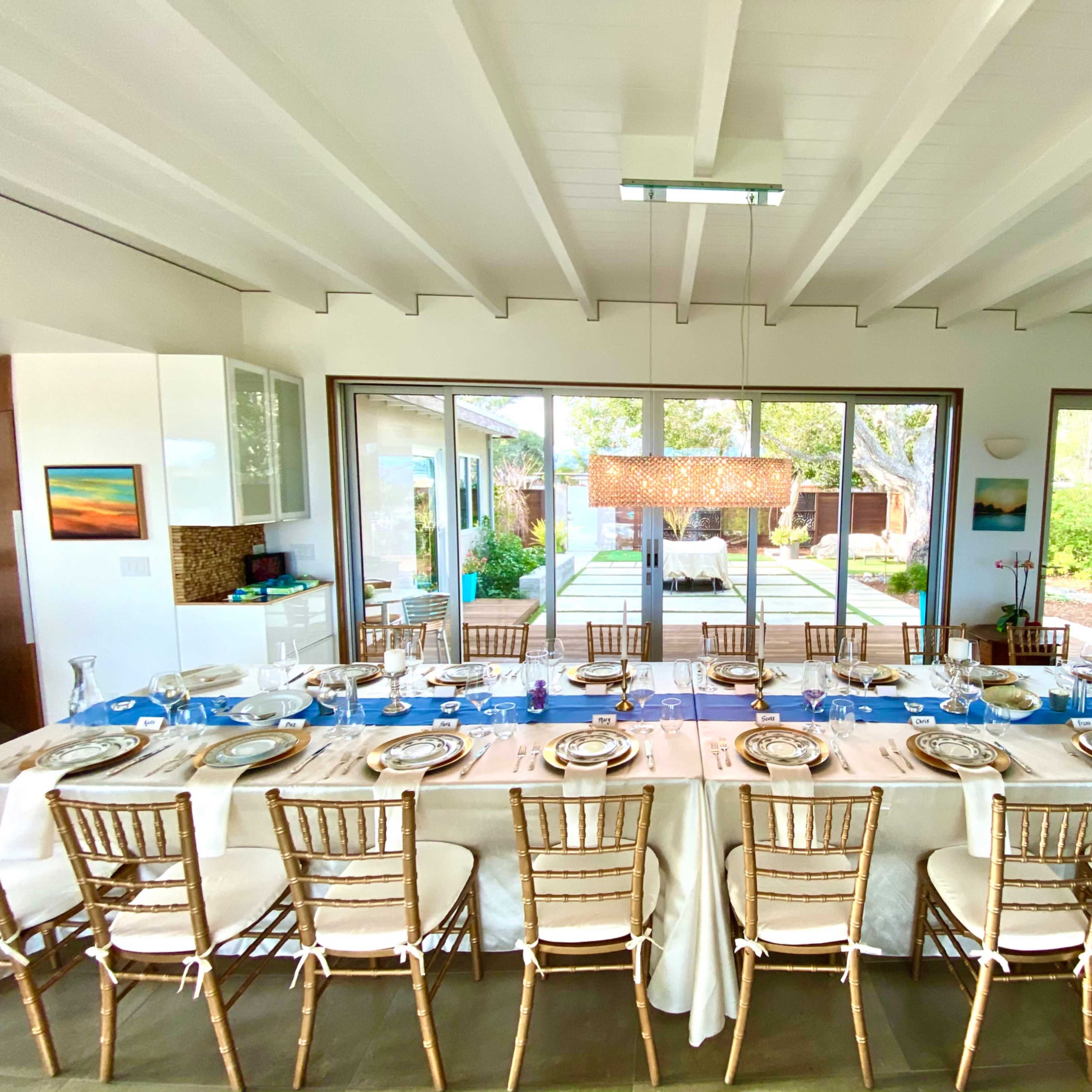 A large dining table is set for a meal in a bright room, with a view of a garden and patio through large glass doors.