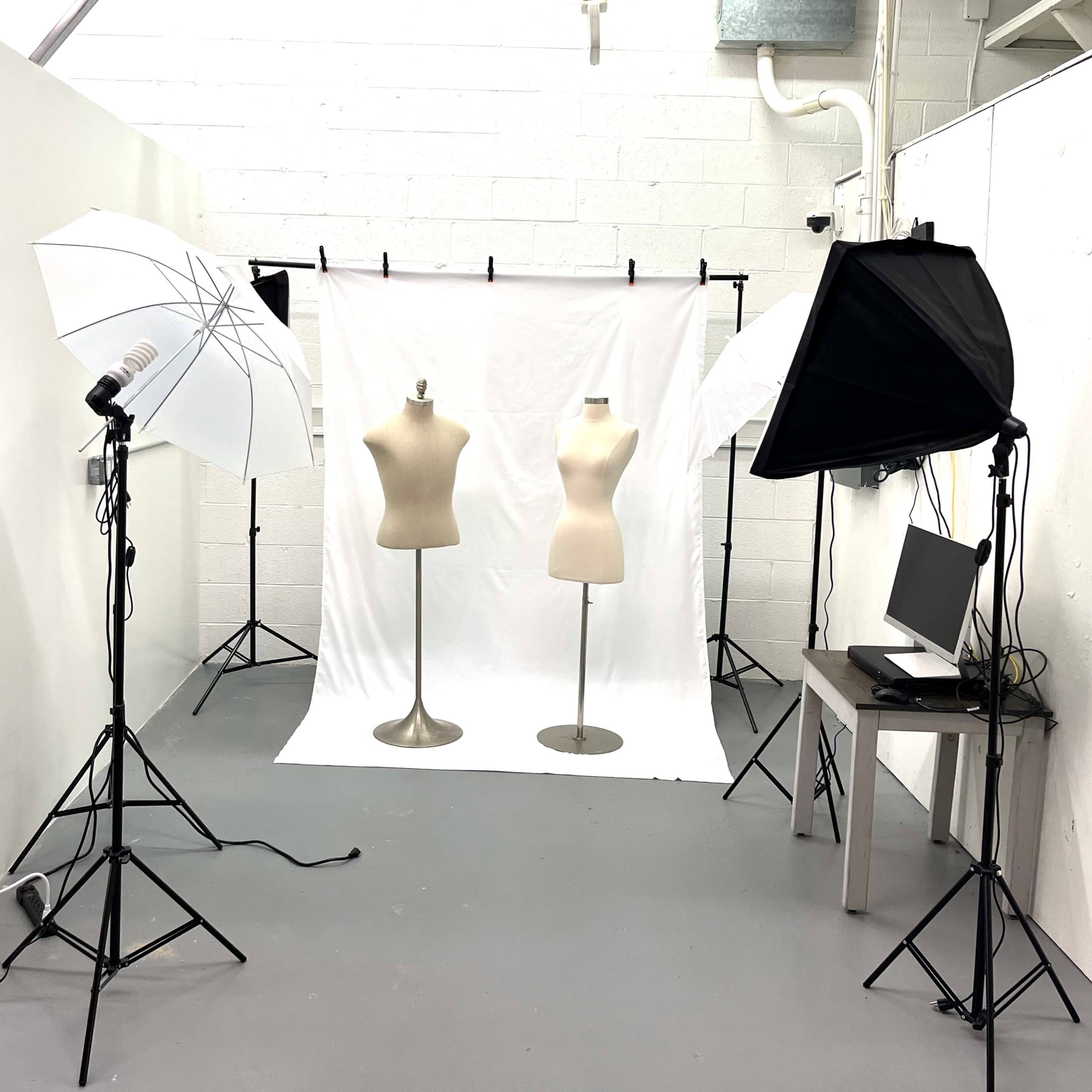 The scene shows a photography setup with two mannequin forms in front of a white backdrop, surrounded by studio lighting equipment and a computer on a table.