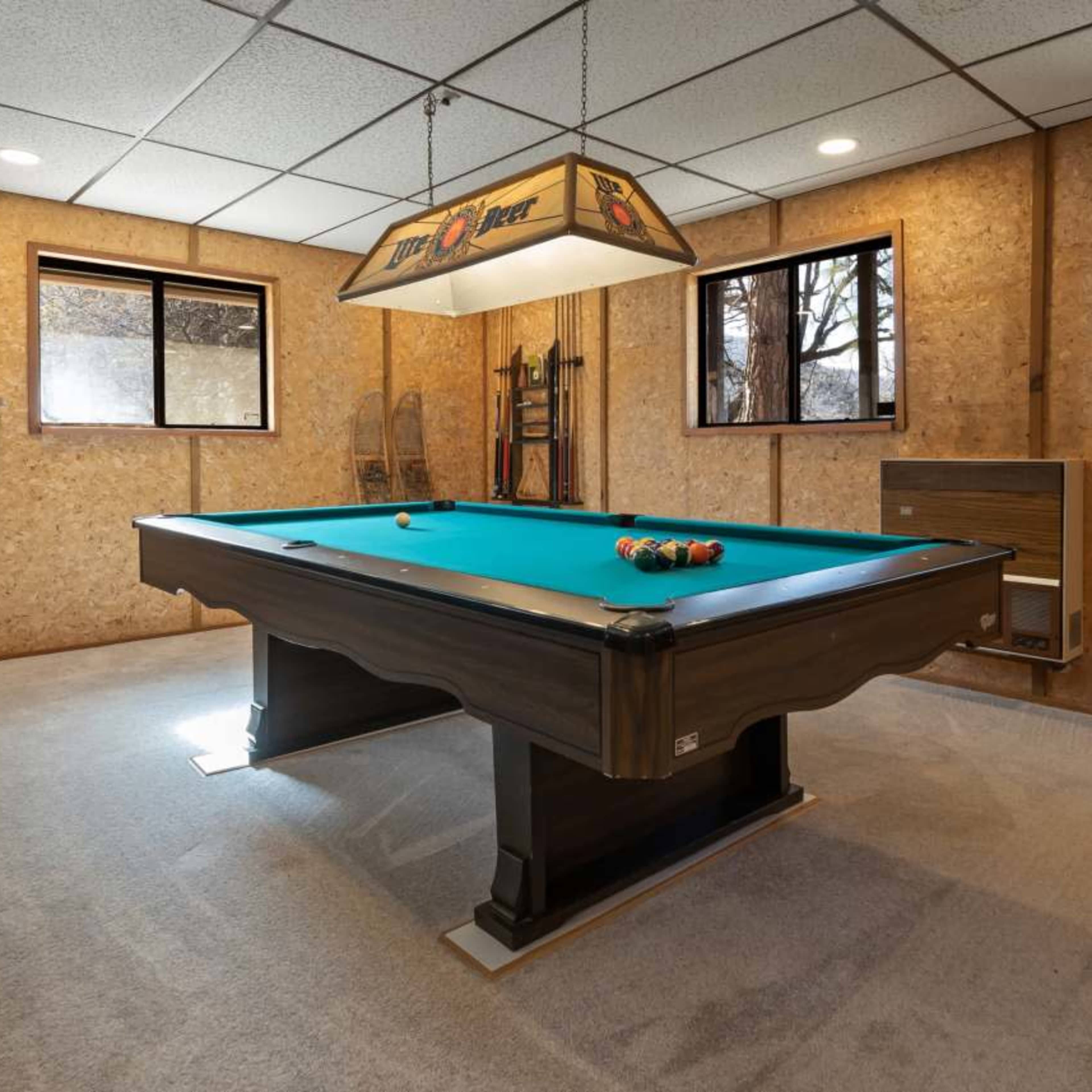 A billiard table with a green felt surface is positioned in a room lined with wood paneling and illuminated by a hanging light fixture.