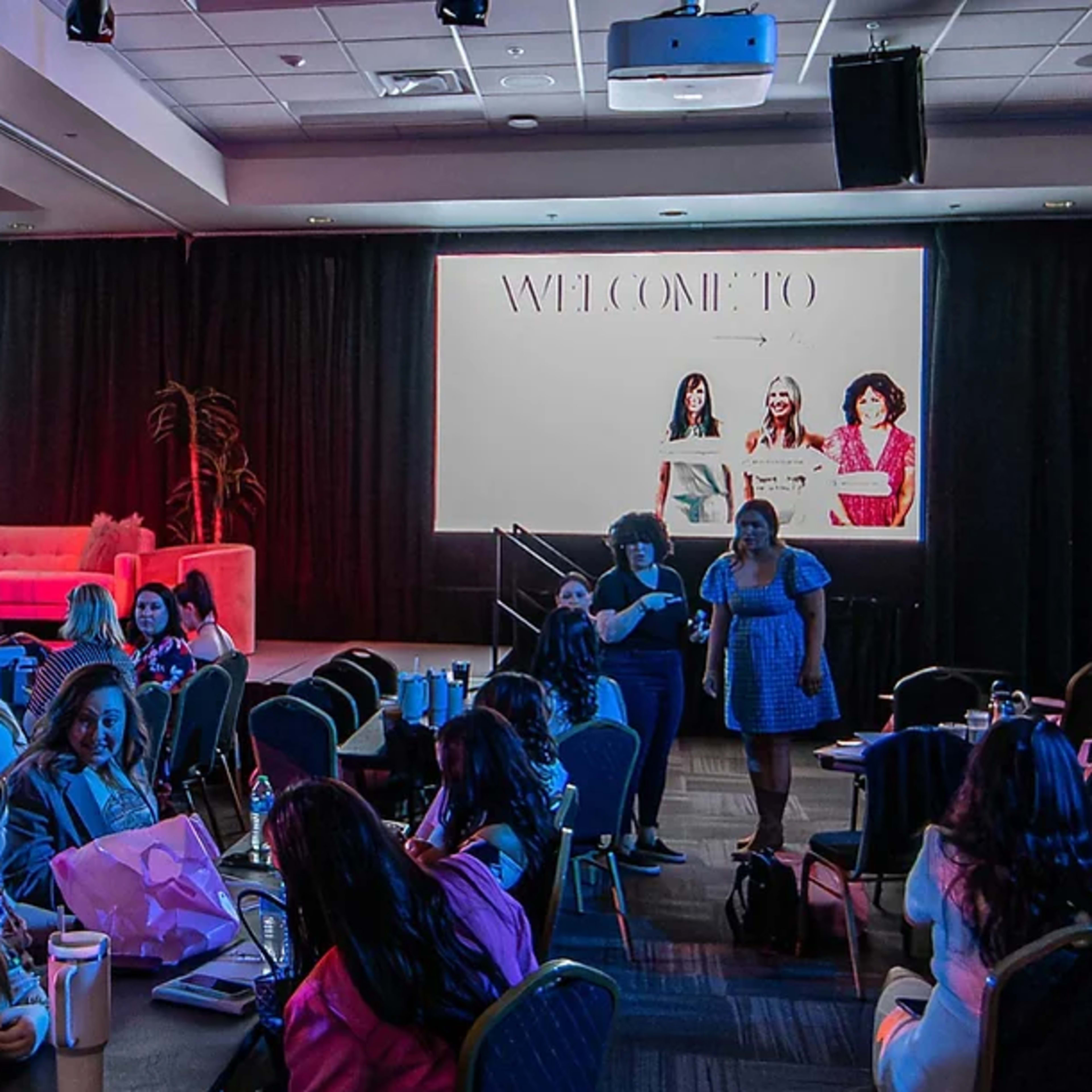 A large conference room filled with participants, seated at tables, facing a stage with a backdrop and a pink couch.