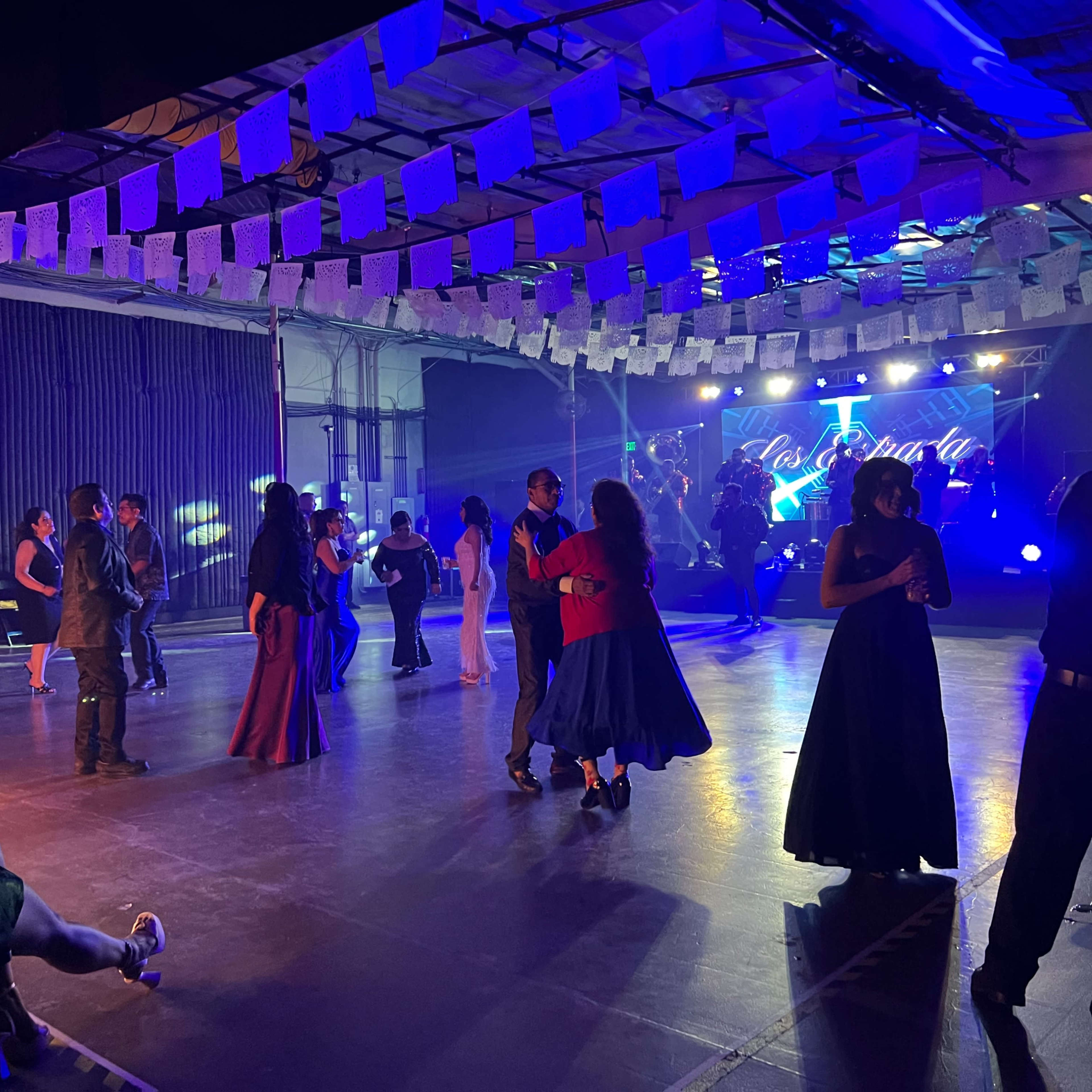 A dance floor is filled with people moving to music under colorful lights, with decorations hanging from the ceiling.