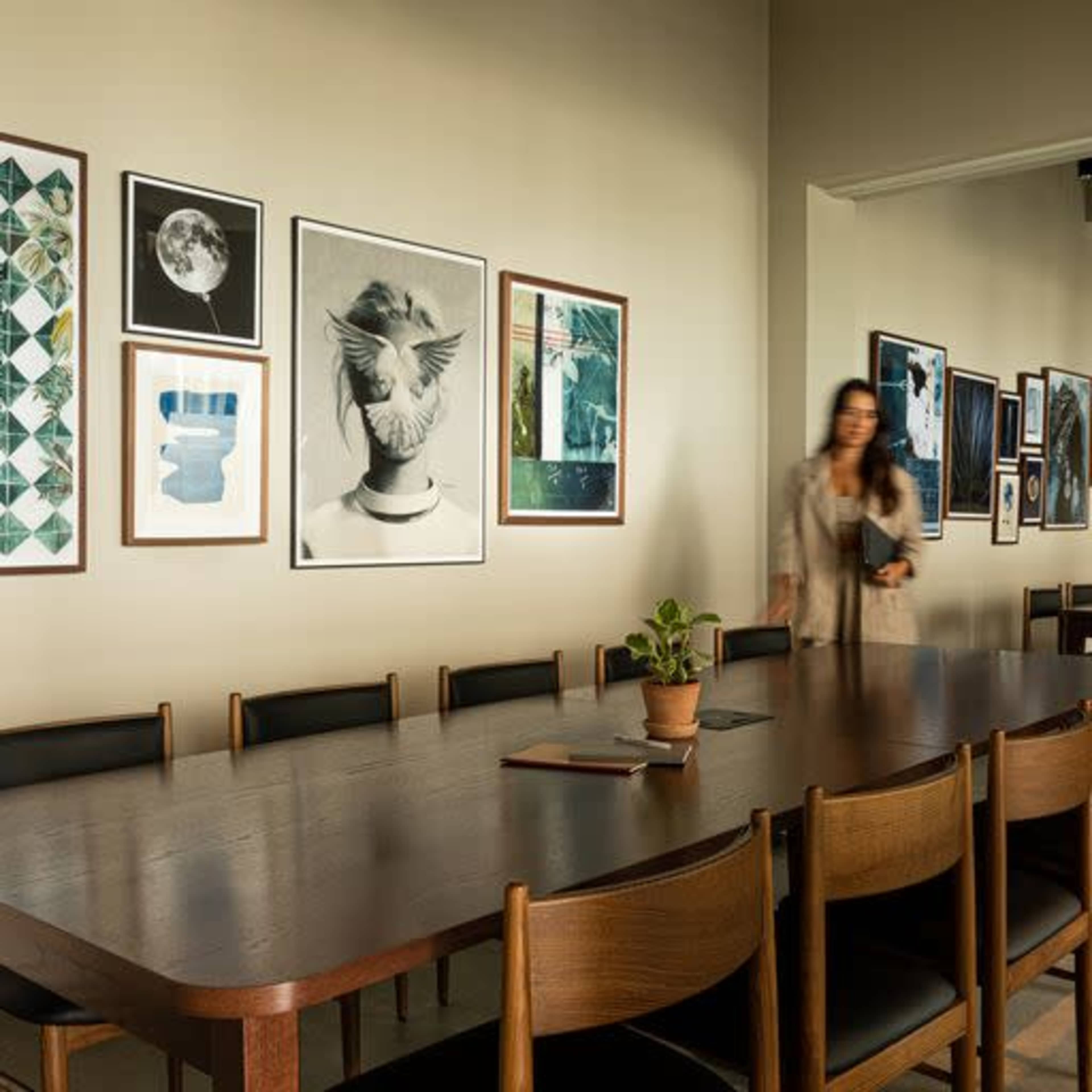 A woman walks past a wooden table set with potted plants, surrounded by chairs, and a wall adorned with various framed artworks.