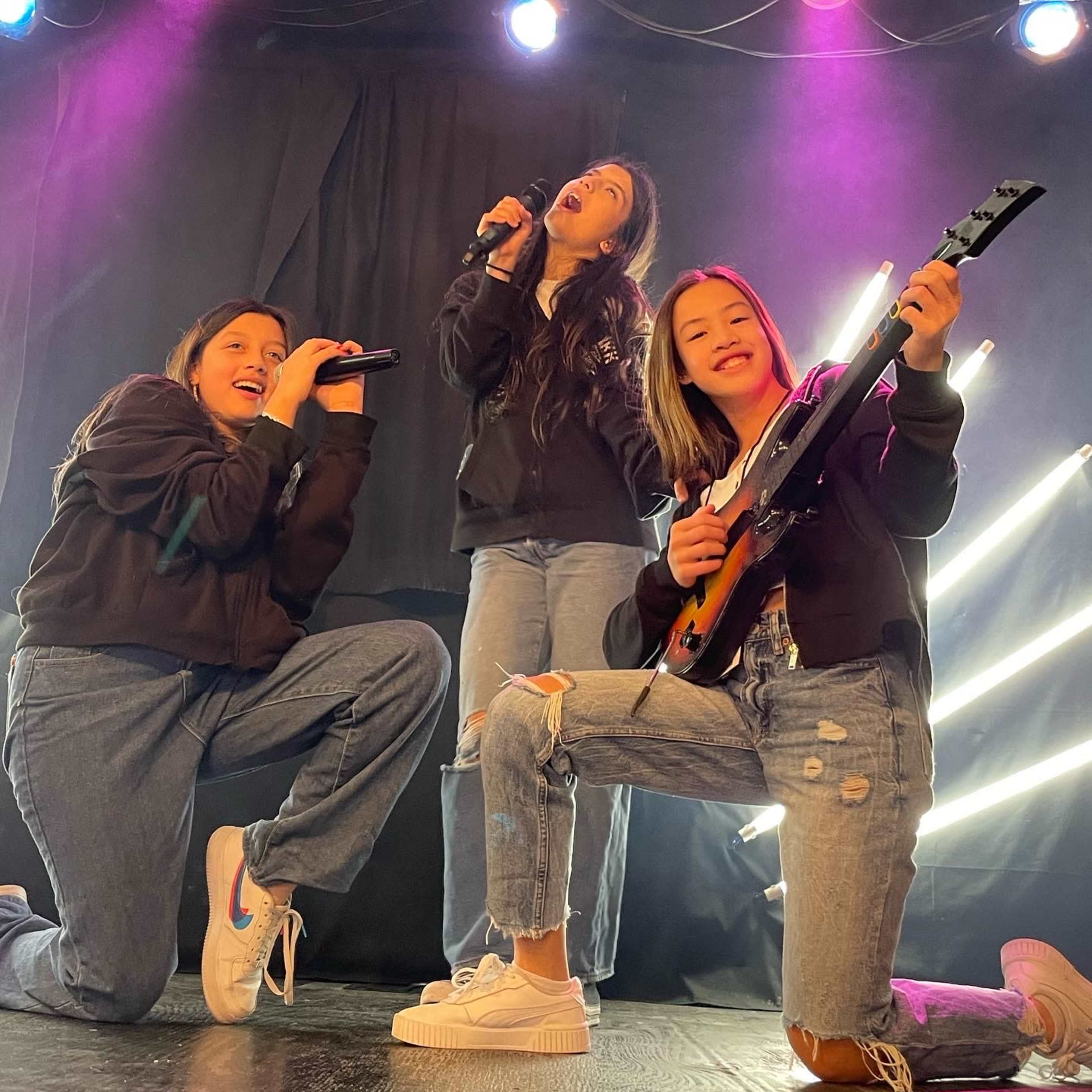 Three individuals perform energetically on stage, with one singing into a microphone and the other two joining with a guitar and microphone, illuminated by colorful stage lights.