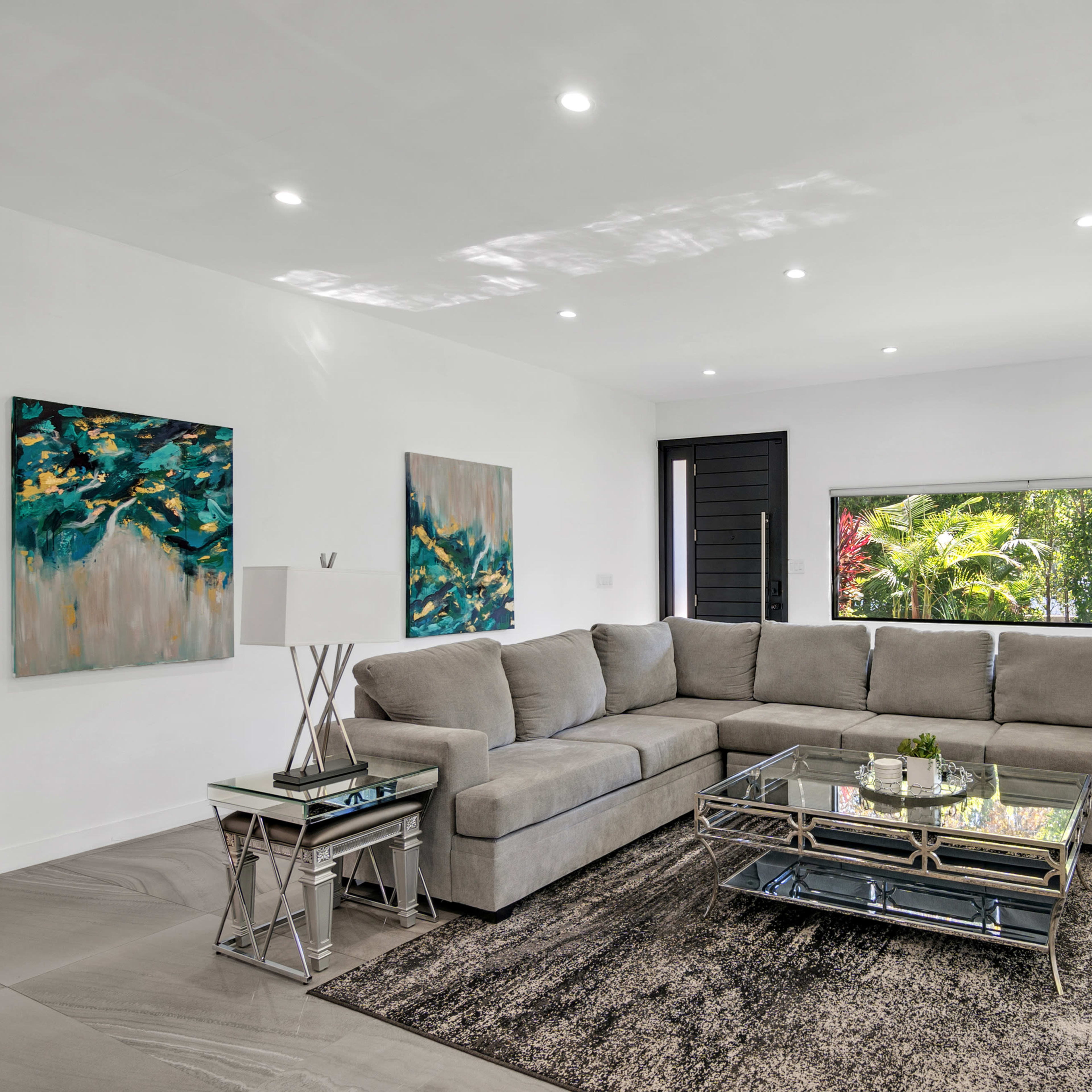 A modern living room features a large gray sectional sofa, a glass coffee table, and two lamps, with abstract paintings on the walls and a window overlooking greenery.