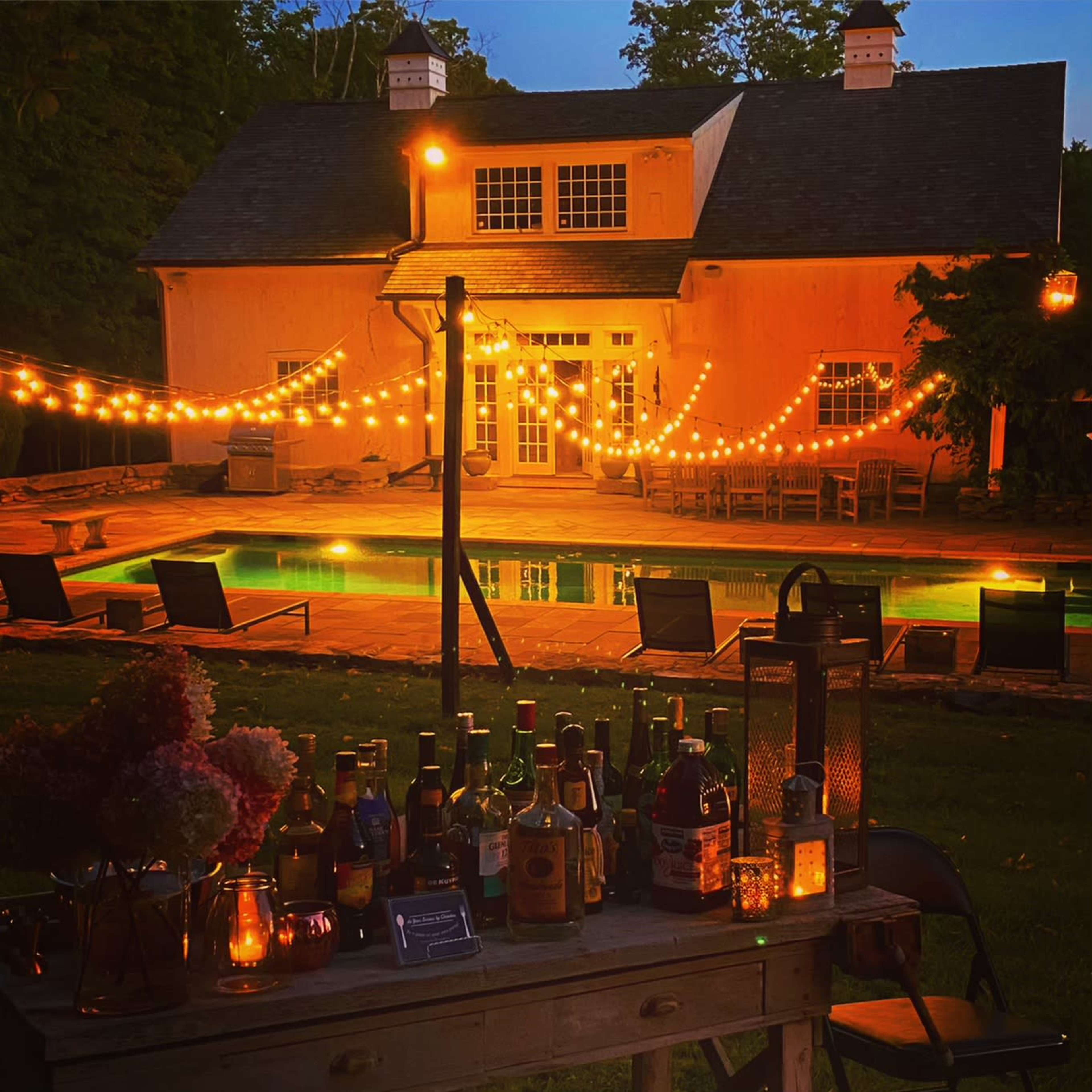 A cozy outdoor setting features a house with string lights illuminated at dusk, a pool in front, and a table displaying various bottles and candles.