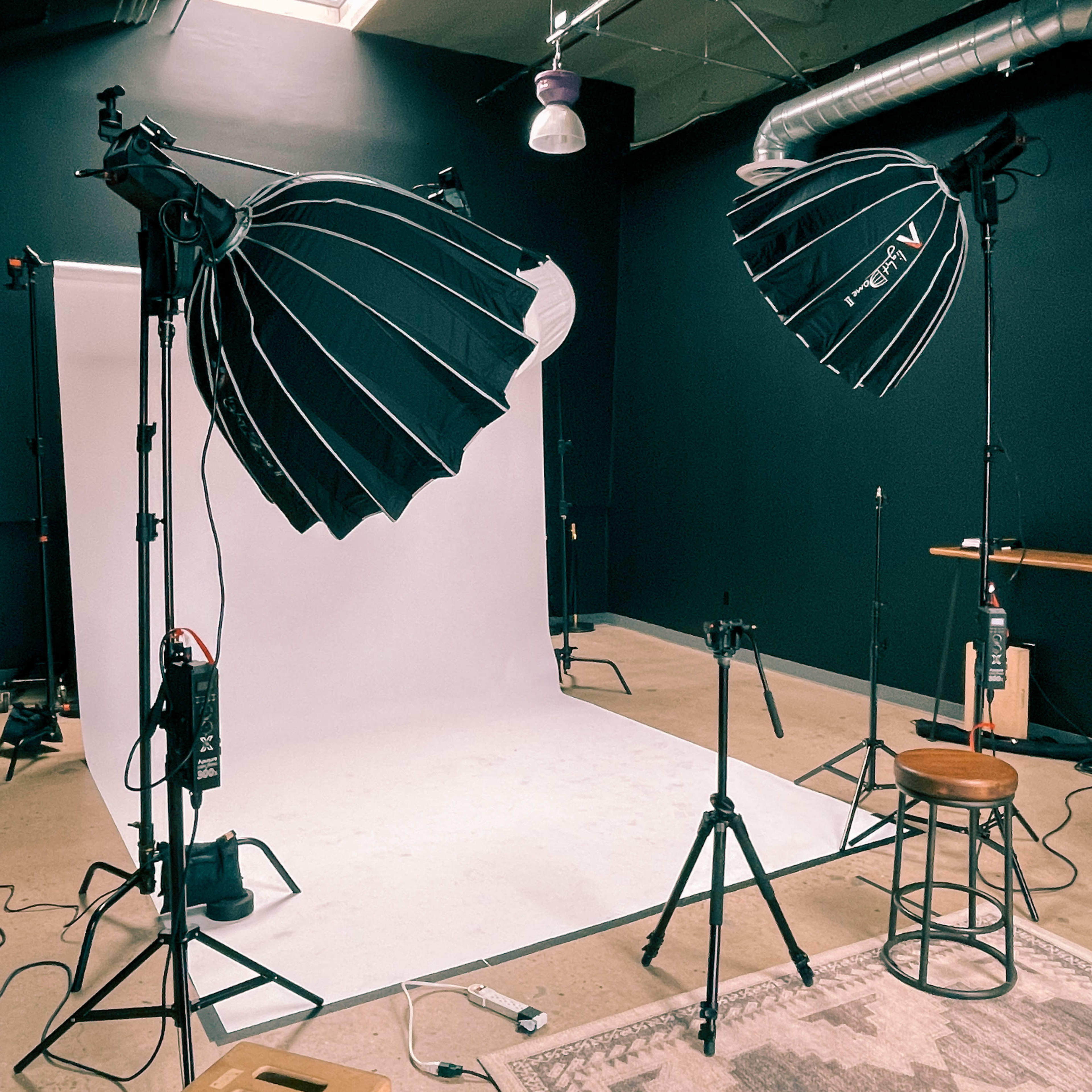 A photography studio setup with two large softbox lights facing a blank white backdrop, a stool, and various equipment on the floor.