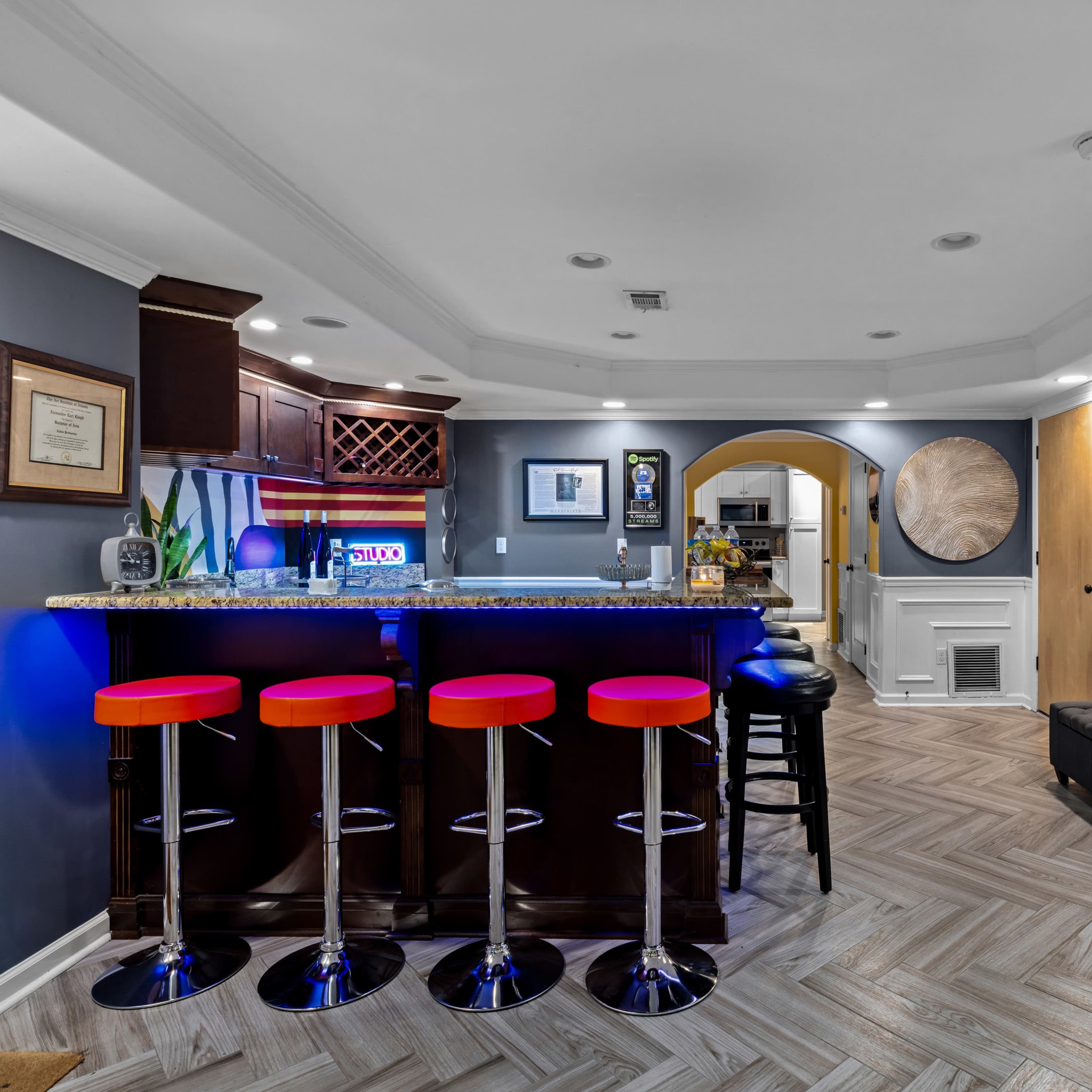 A modern basement bar area featuring four red bar stools, dark cabinetry, and an artistic wall display.
