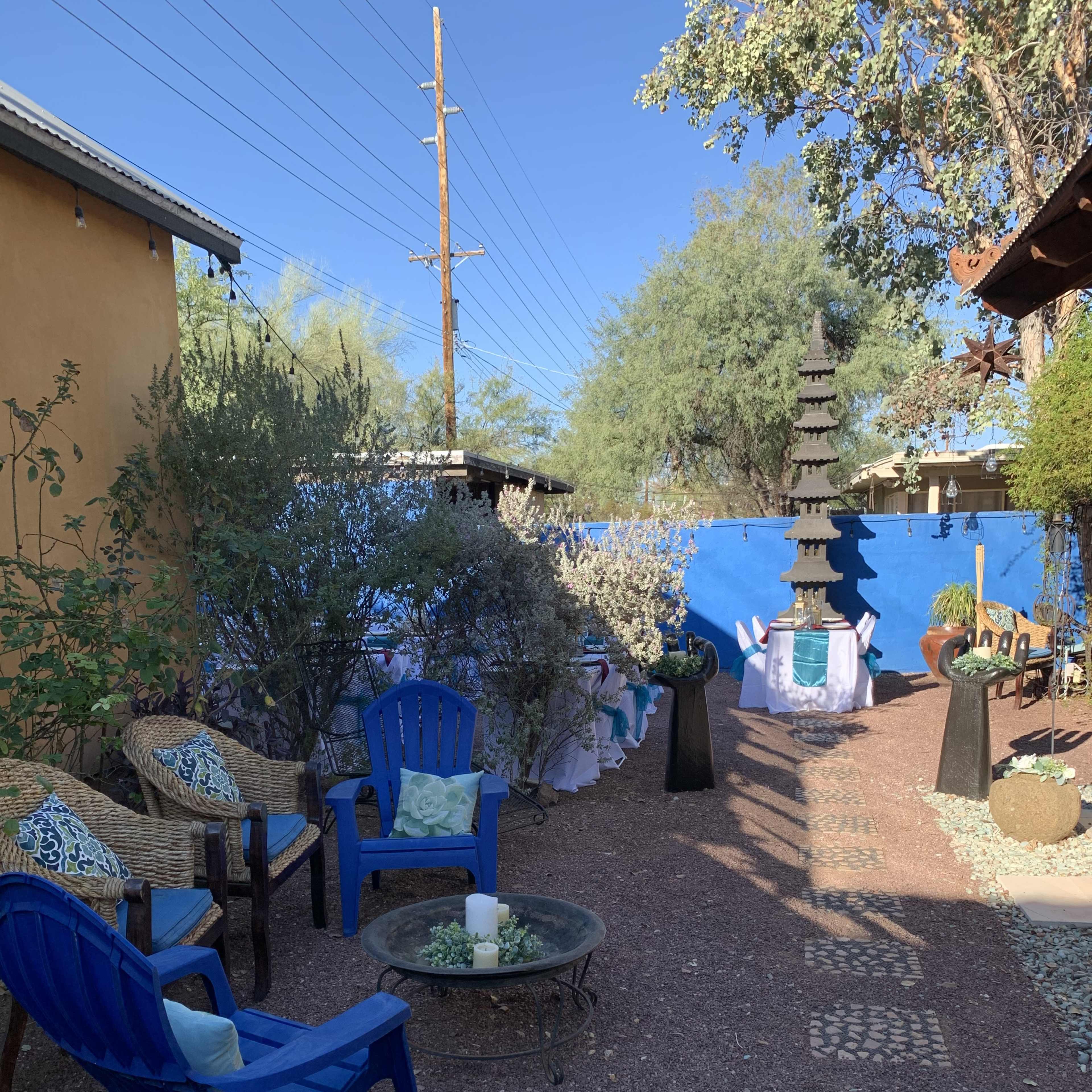 A landscaped outdoor space with blue chairs, a circular table, pathway stones, a decorative tower, and various plants against a blue wall.