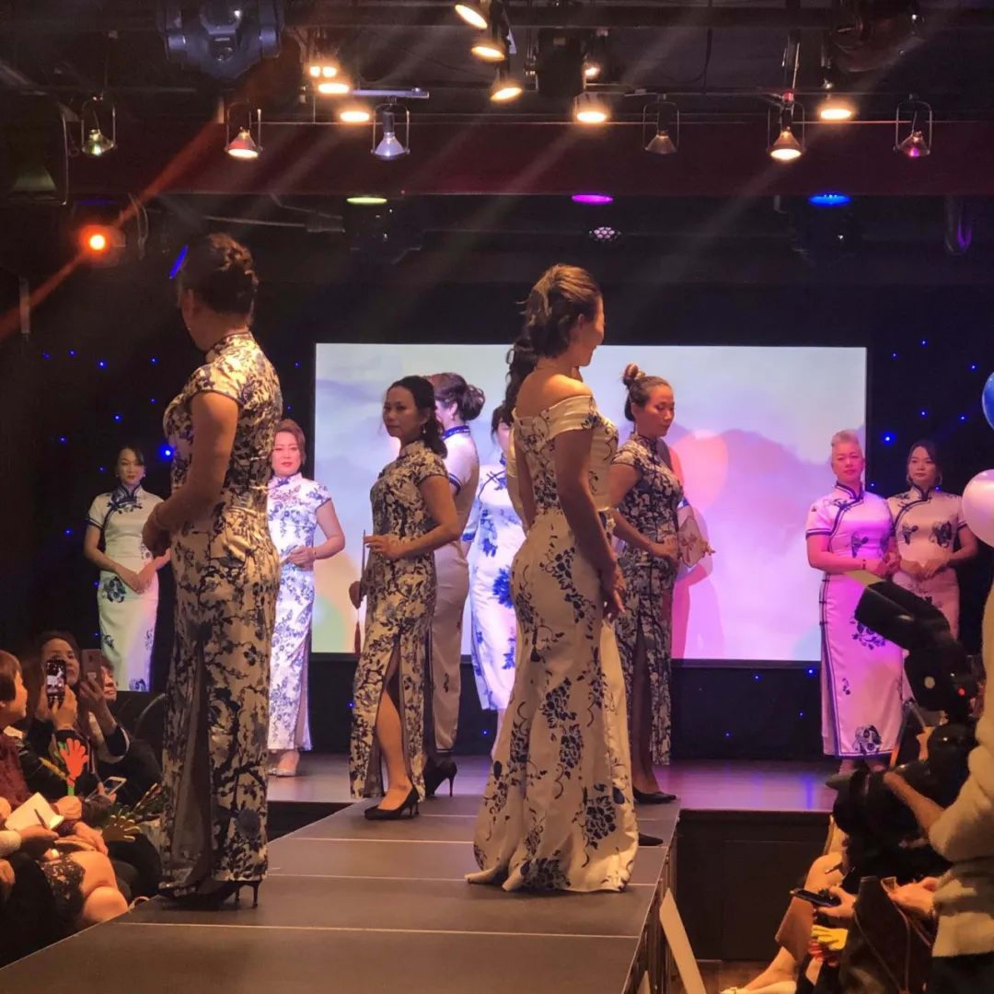 Models wearing traditional dresses walk down a runway while an audience observes.