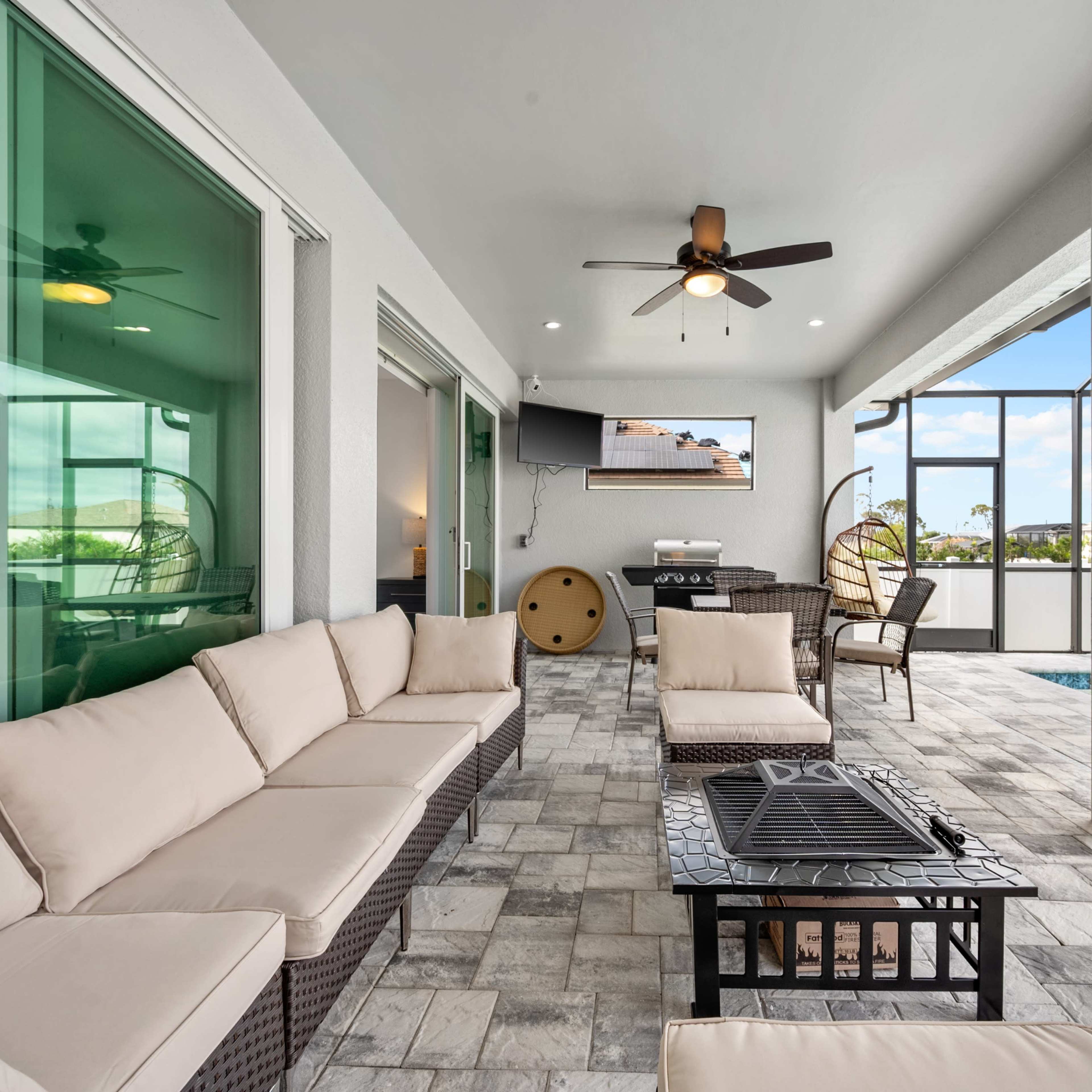 A modern outdoor patio with a sectional seating arrangement, a fire pit, and a television mounted on the wall, overlooking a pool area enclosed by a screen.
